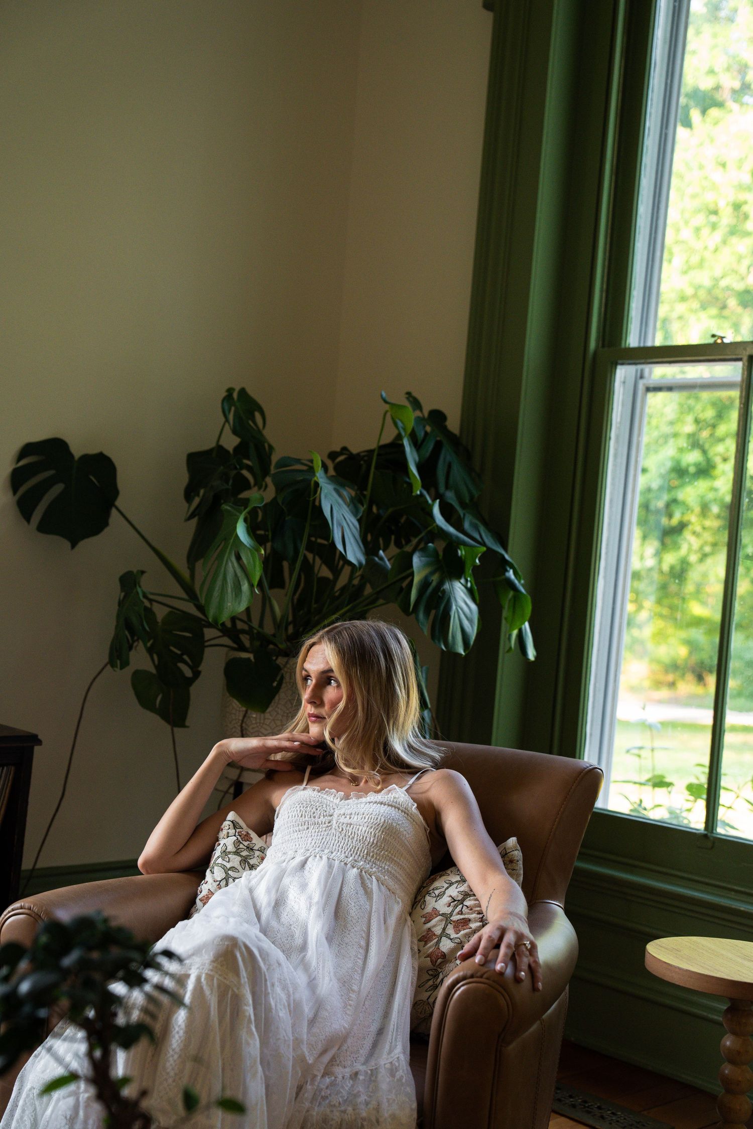 Bride in white sundress sits thoughtfully in a leather armchair beside a green-trimmed window with plants.