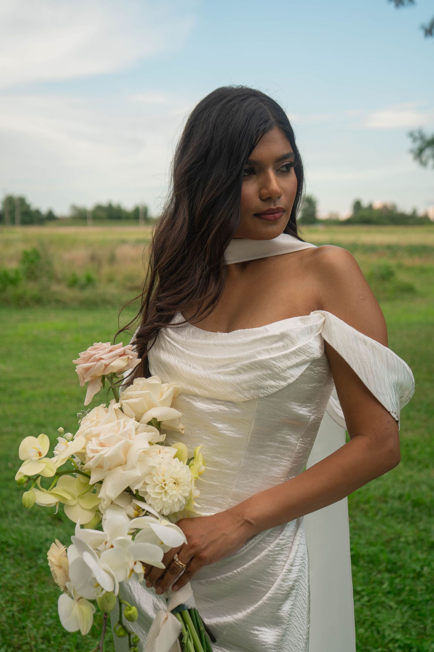 Dark-haired bride in white off-shoulder dress holds cascading bouquet of white orchids and blush roses outdoors.