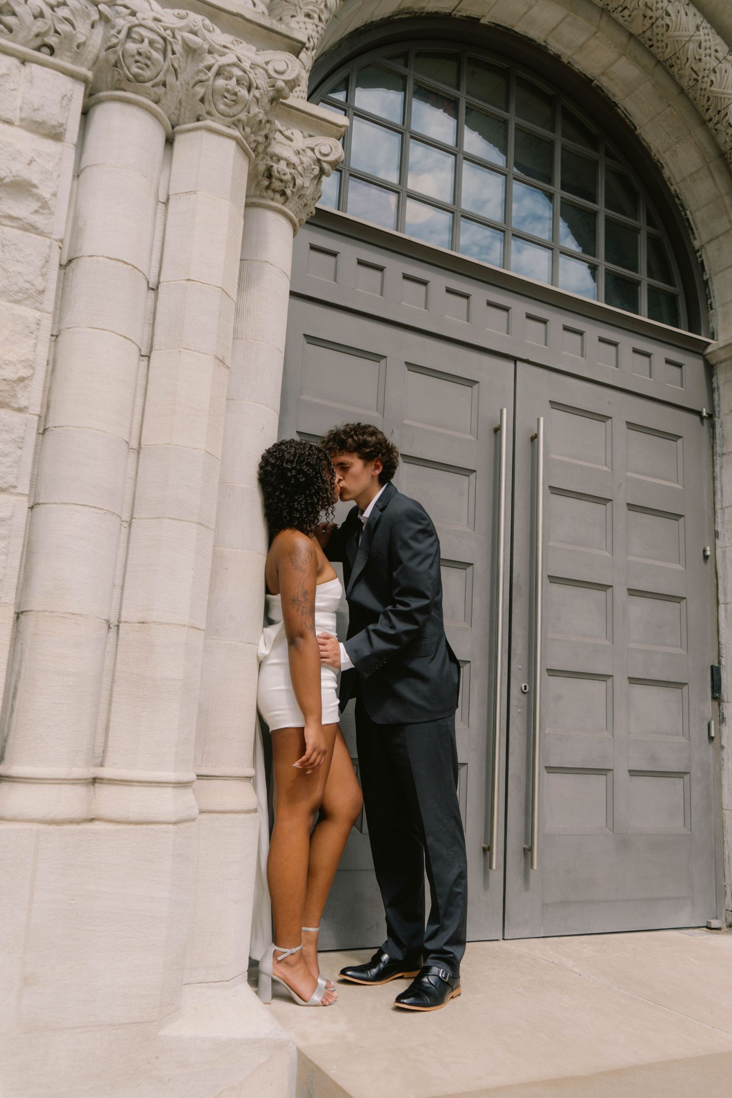 Bride in white mini dress and groom in dark suit sharing an intimate moment by ornate stone columns.