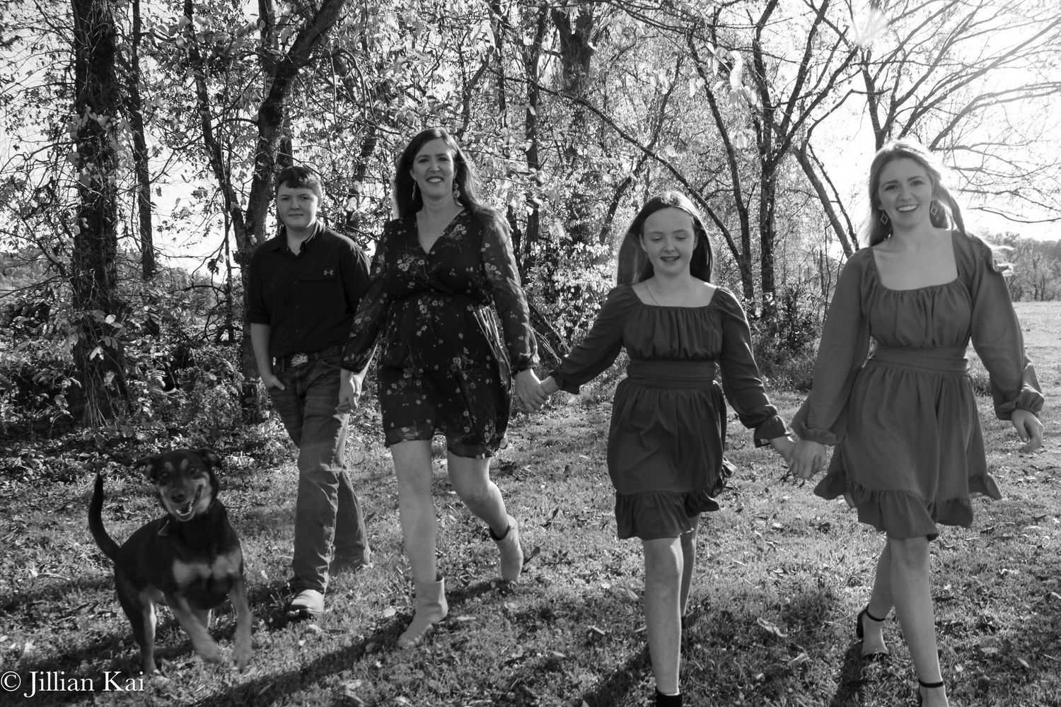 Black and white photo of family walking with small dog through wooded area holding hands.