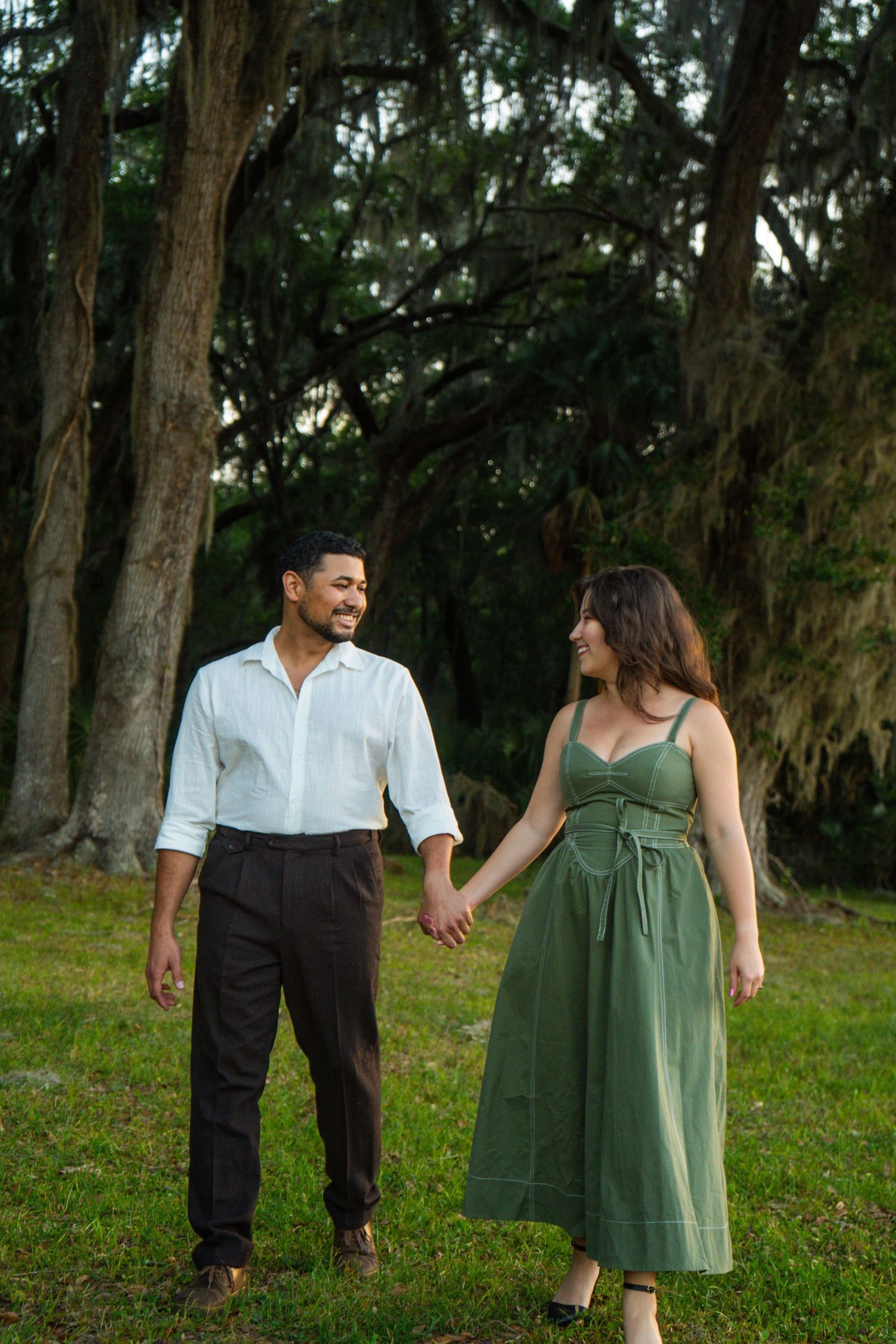 Smiling couple holding hands walking in forest with Spanish moss trees.