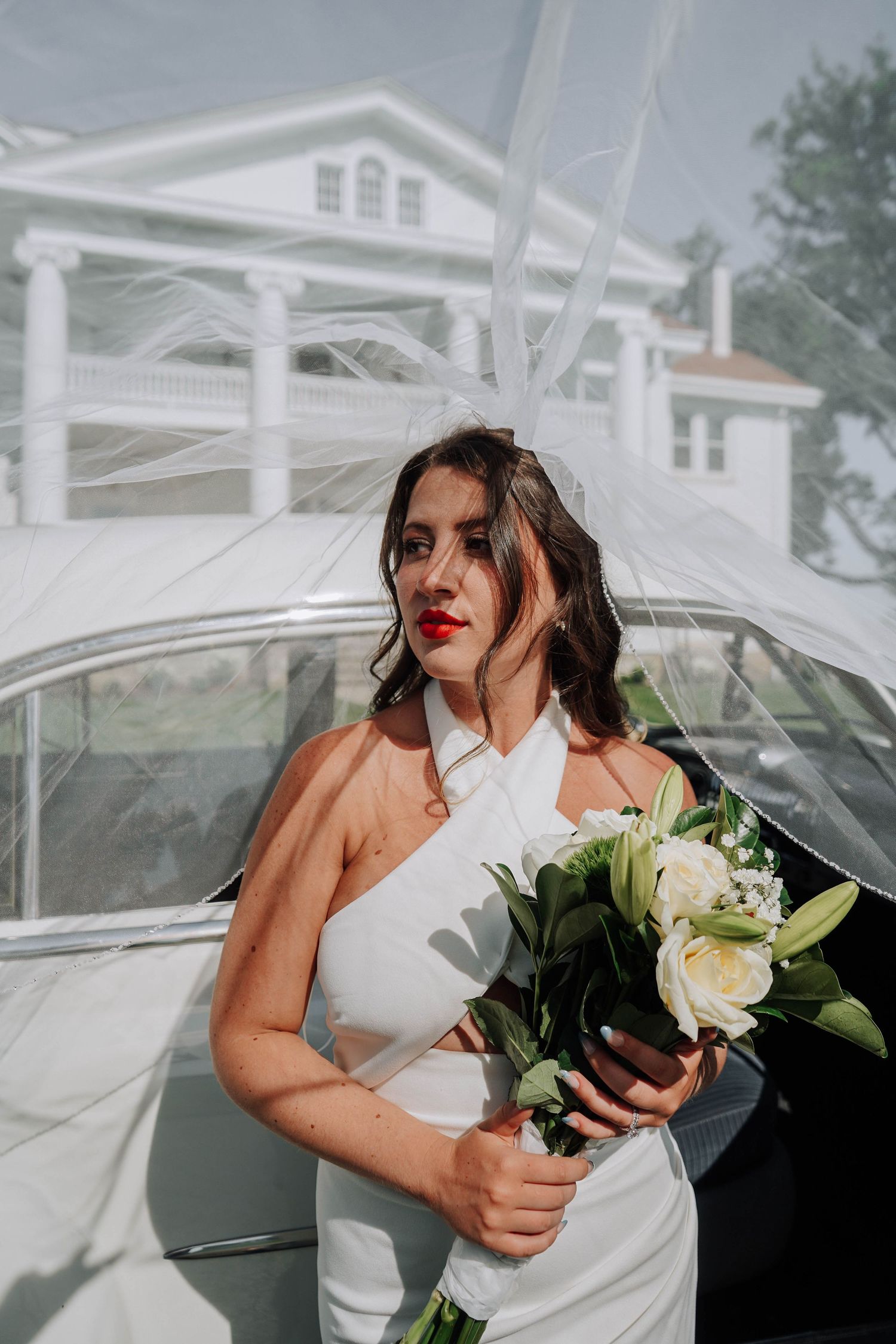 Bride in white halter gown holding bouquet of white lilies and roses beside vintage car with veil flowing.