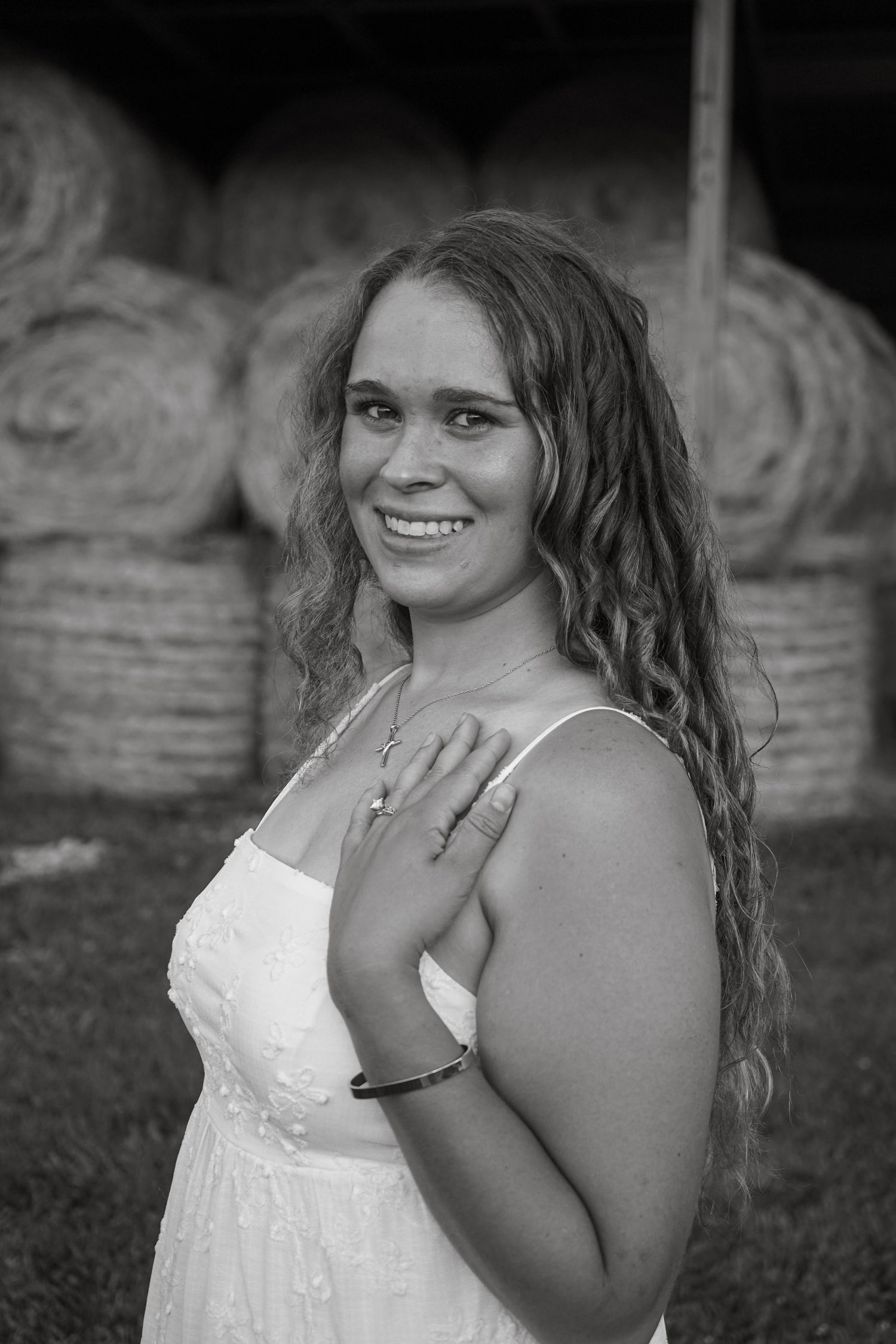 Young woman in white dress smiling in front of hay bales in black and white photo.