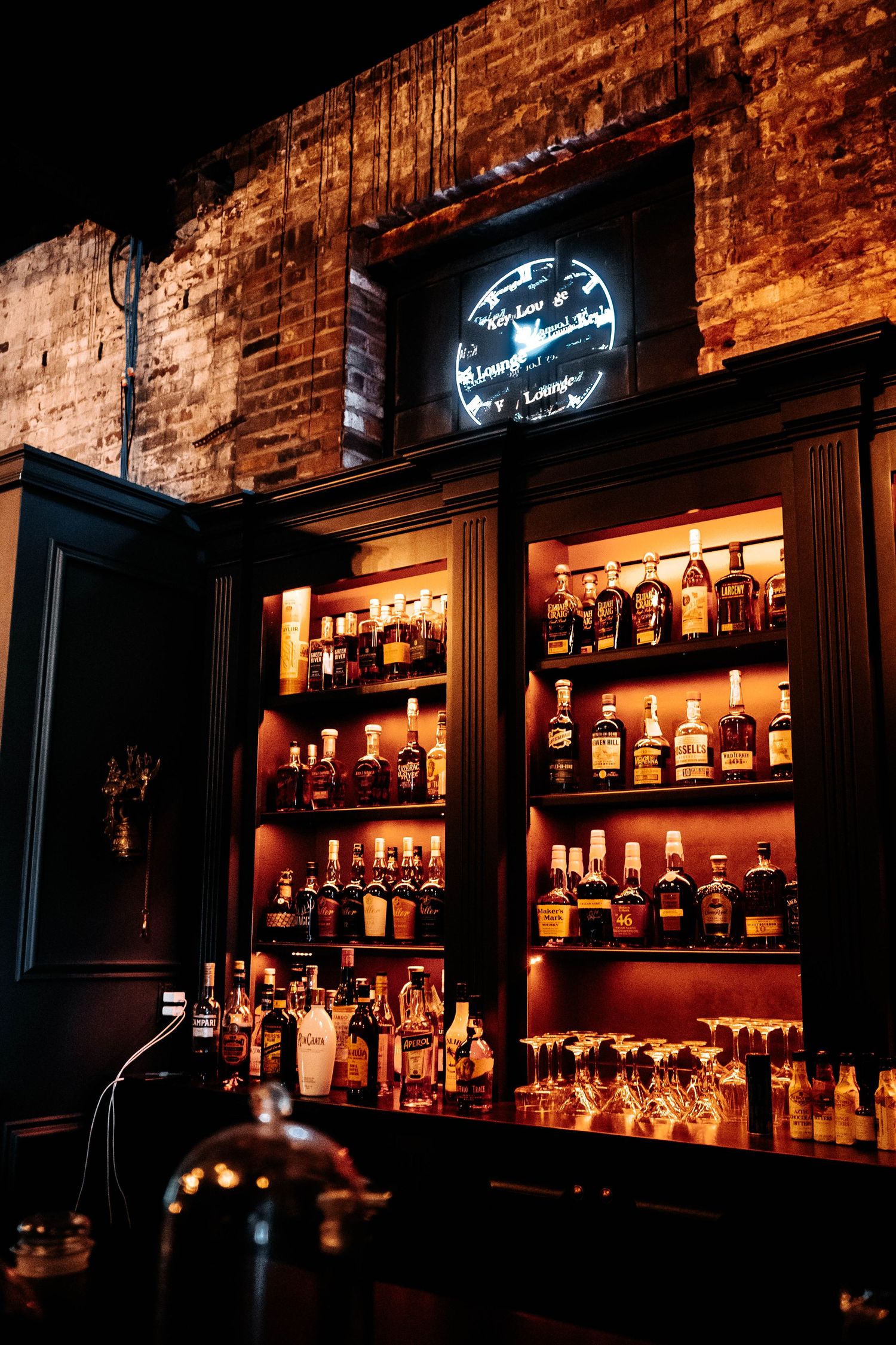 Warmly lit bar shelves displaying whiskey bottles against exposed brick wall with neon lounge sign above.