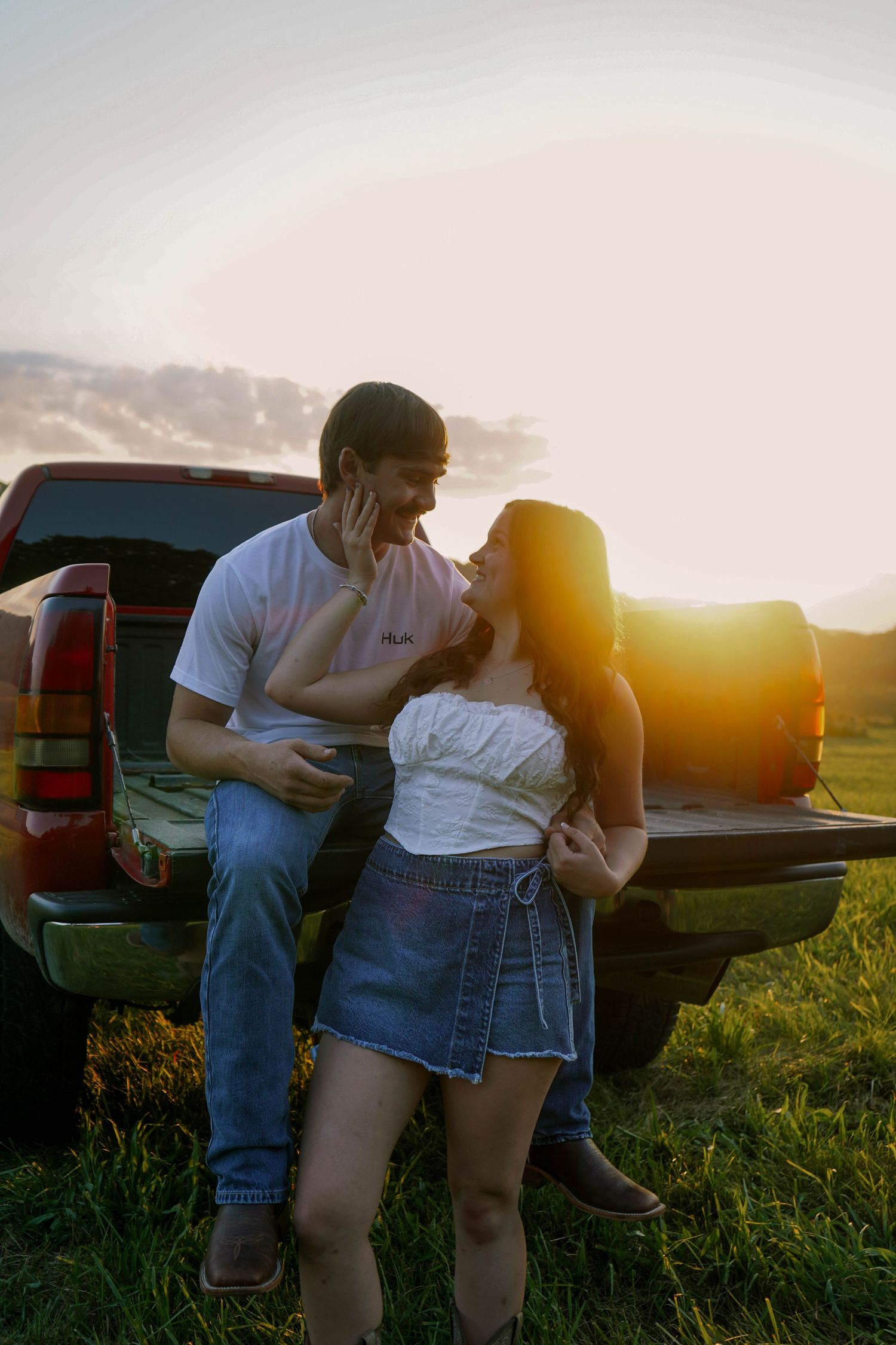 Laughing couple sitting on the tailgate of a red pickup truck during a warm glowing sunset in a rural field.