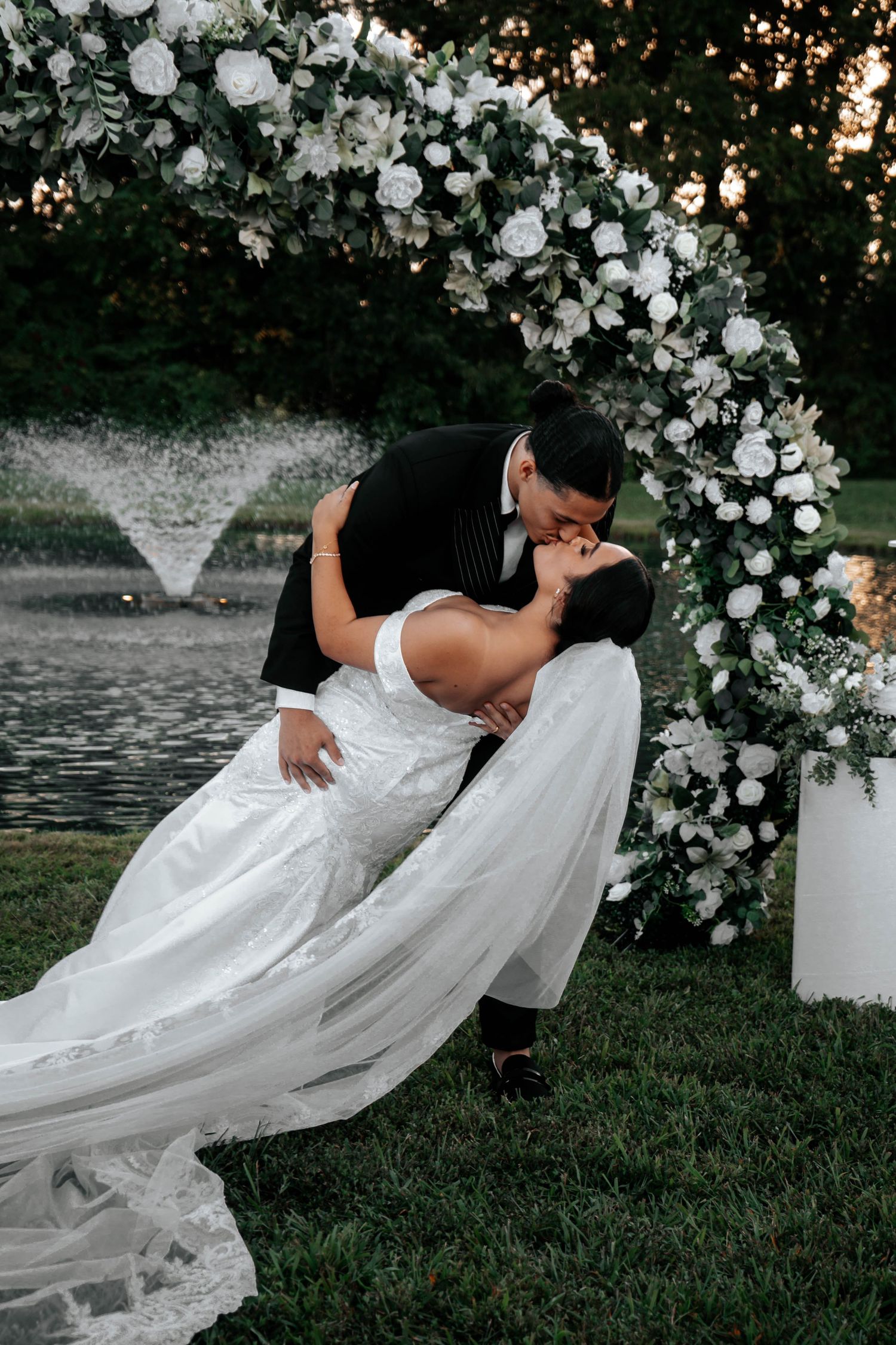 Groom dips bride in romantic kiss beside white rose floral arch with fountain backdrop at sunset.