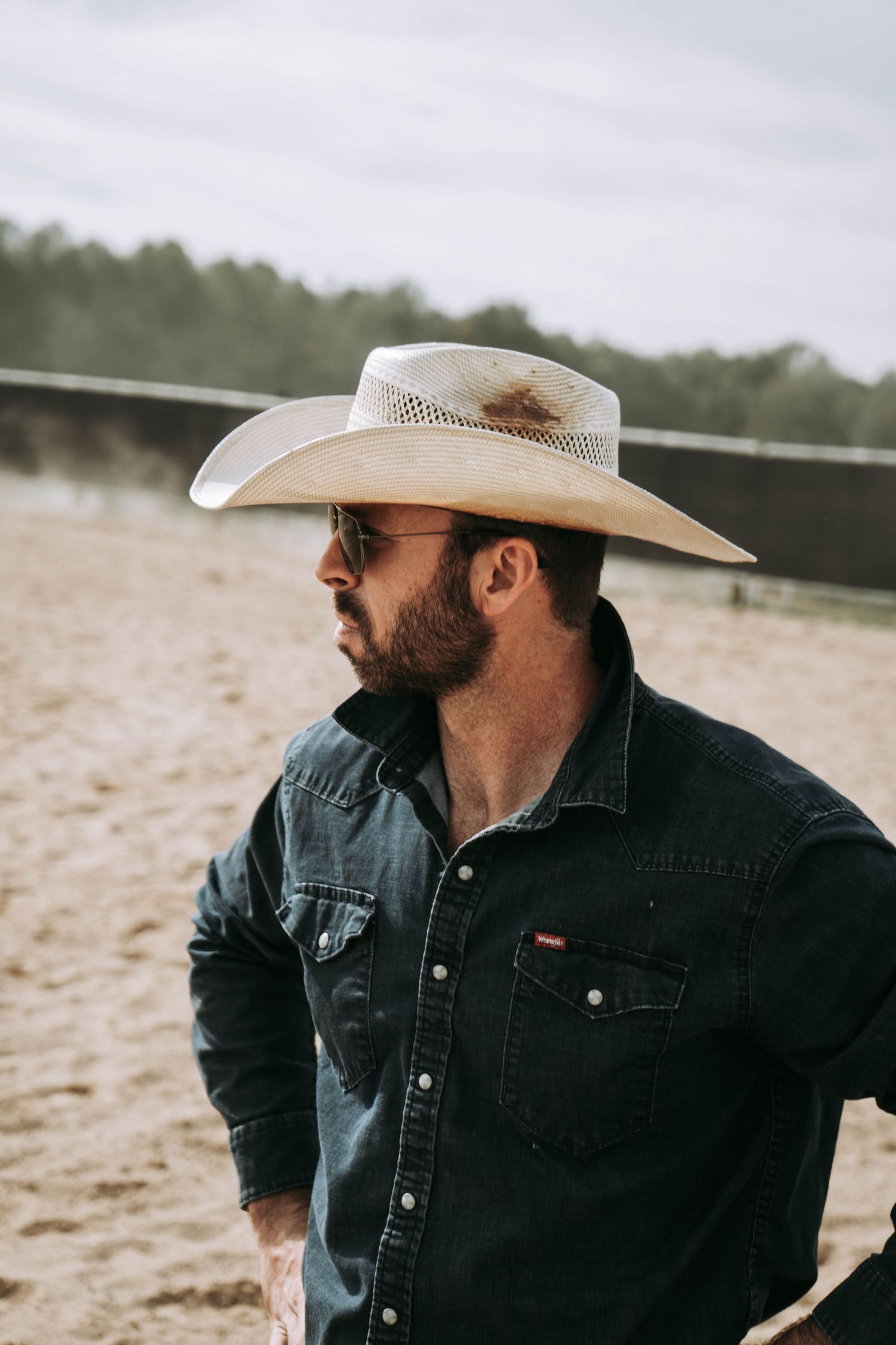 Bearded man wearing a cowboy hat and sunglasses in a dark denim shirt at an outdoor sandy arena.