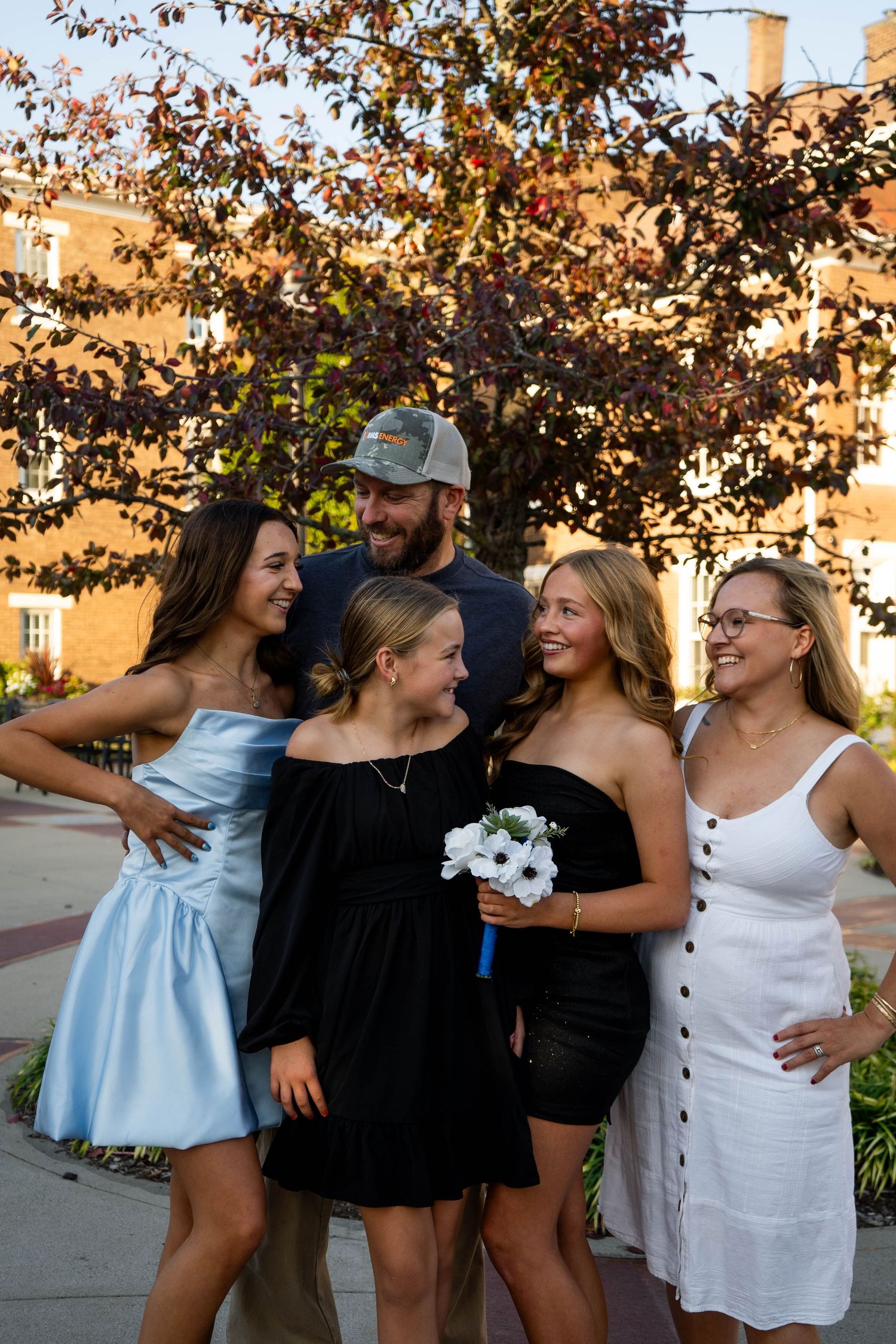 A happy family of five laughs together outdoors beneath a decorative tree during a prom photo session.