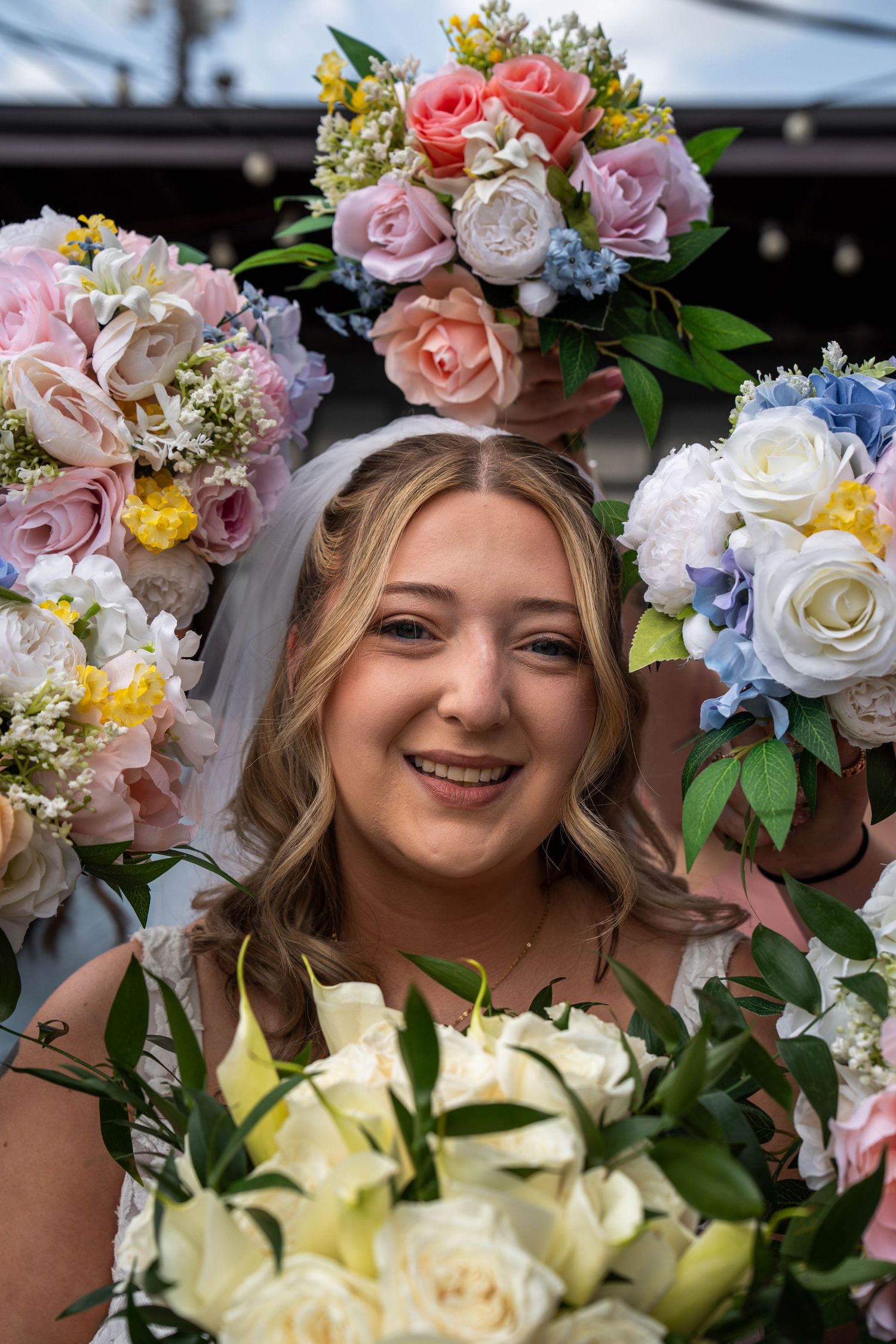 Smiling bride surrounded by colorful pink, white, and blue floral bouquets on her wedding day.