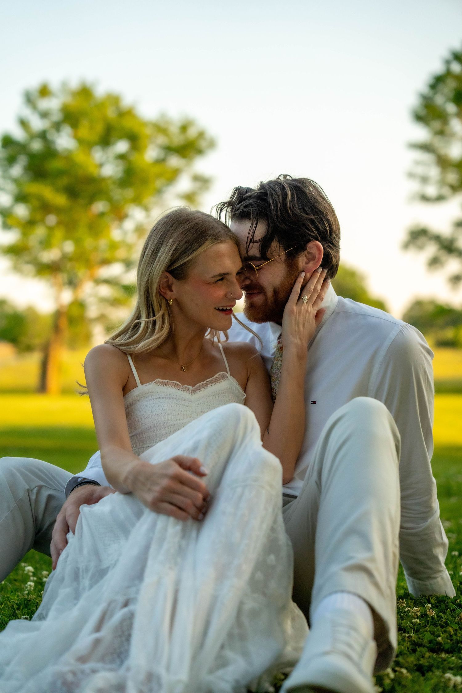 Couple sitting on grass in golden hour light, woman in white dress smiling as man leans in close romantically.