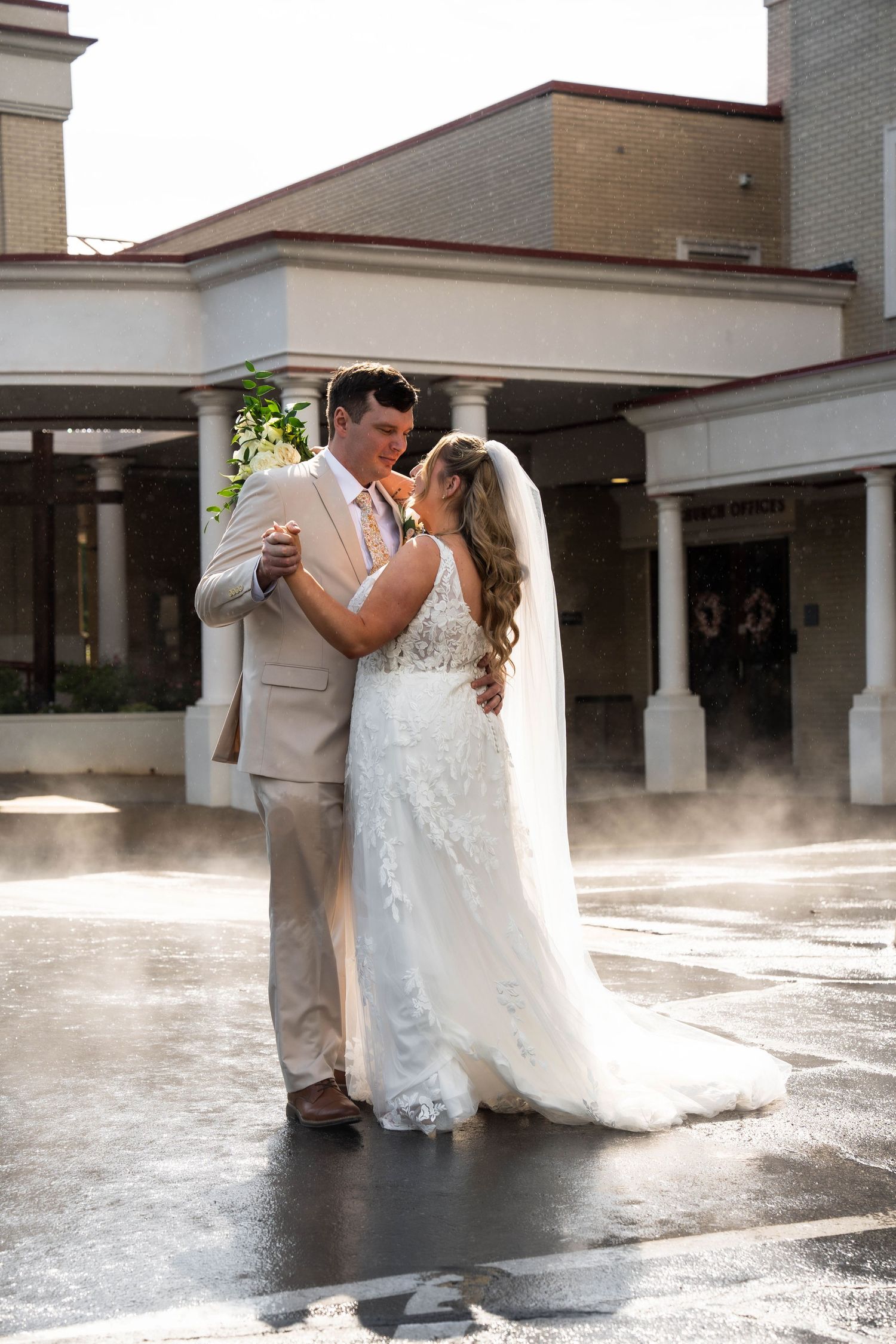 Bride and groom share a romantic dance outdoors in misty light, she in lace gown, he in tan suit.