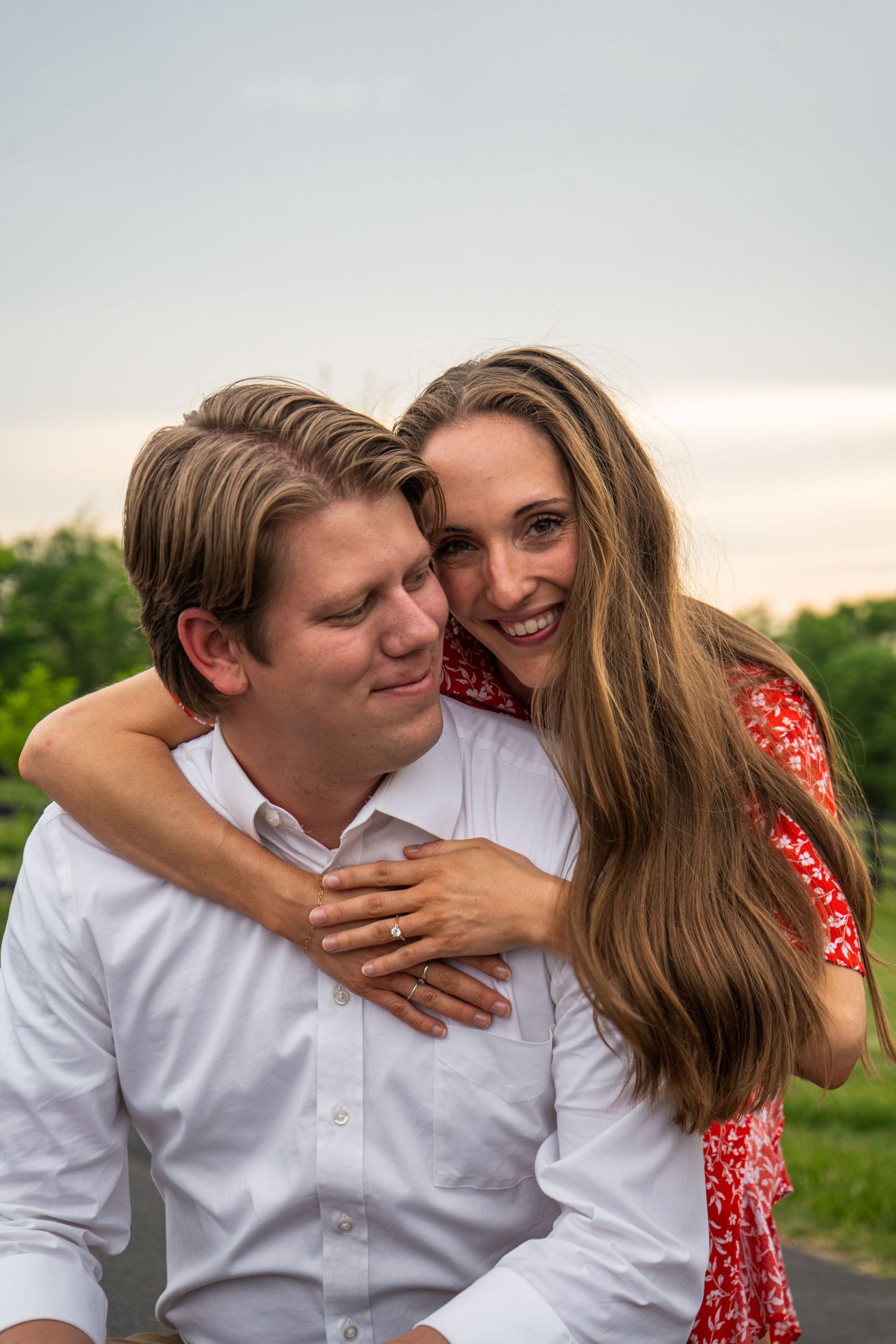 Woman in red floral dress hugging man from behind, both smiling warmly at sunset outdoors.