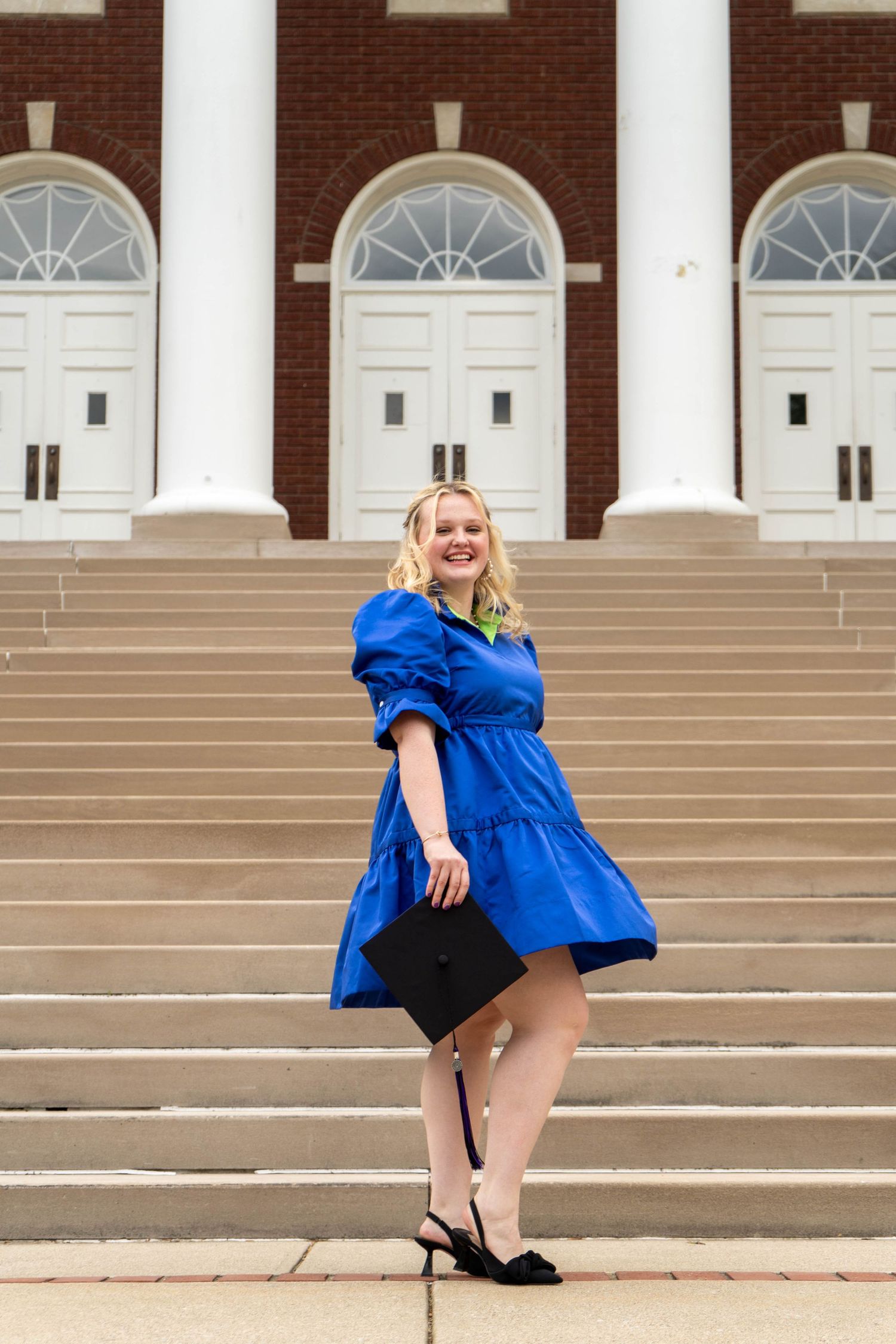 Smiling blonde graduate in blue dress holding mortarboard on university steps.