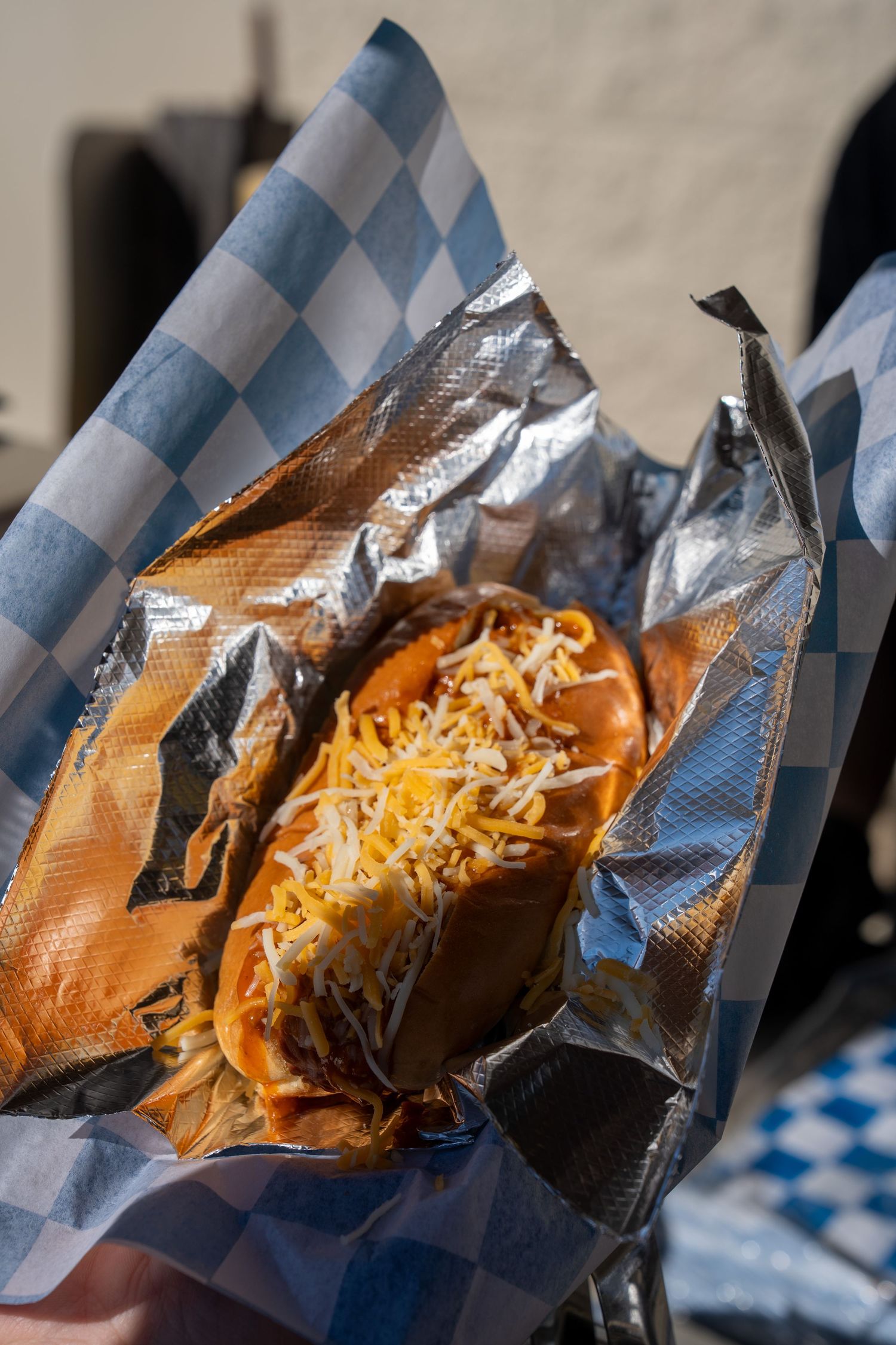 A chili cheese hot dog wrapped in foil and served in a blue checkered paper basket.
