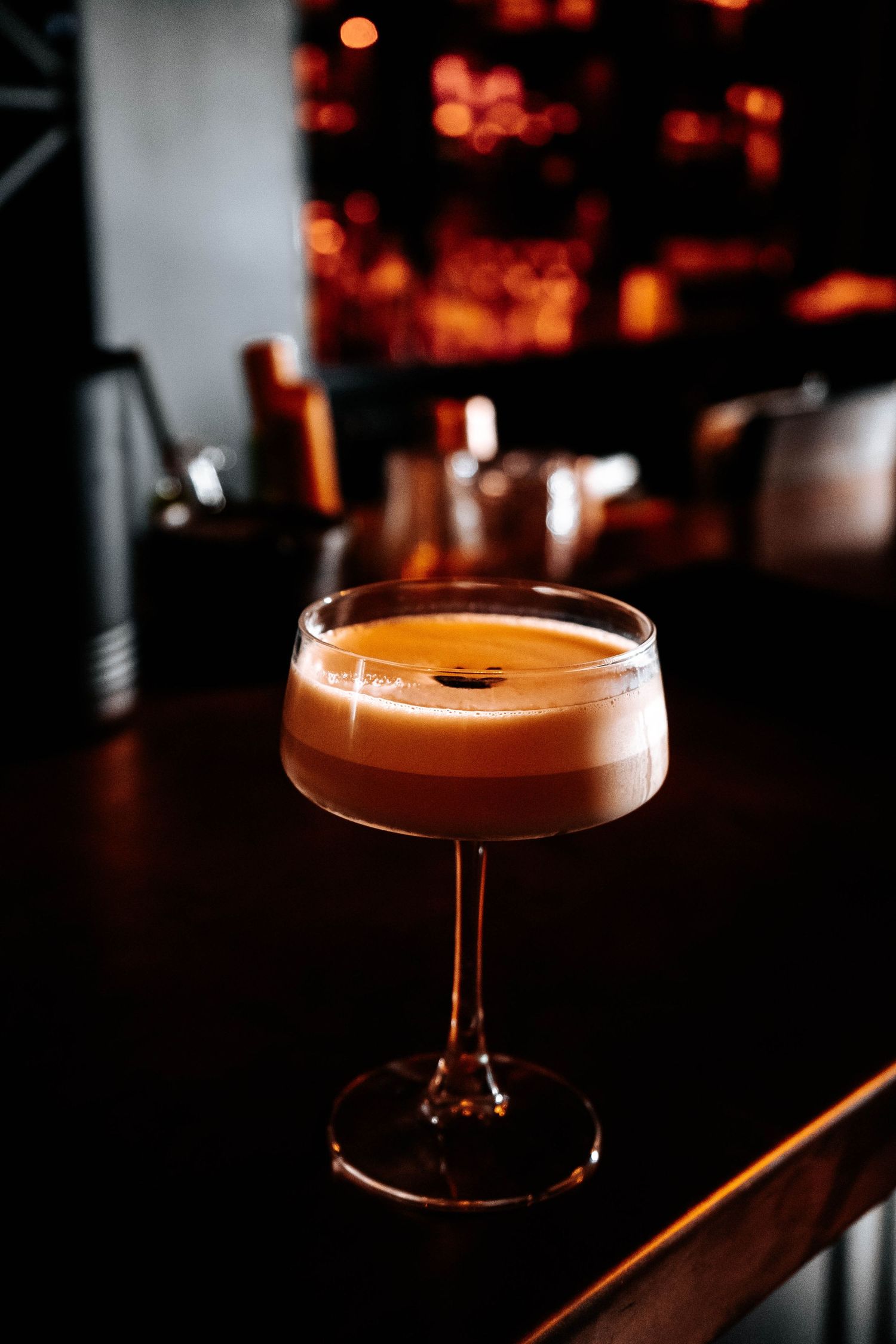 An espresso martini cocktail in a coupe glass on a dark bar counter with warm bokeh lights in the background.