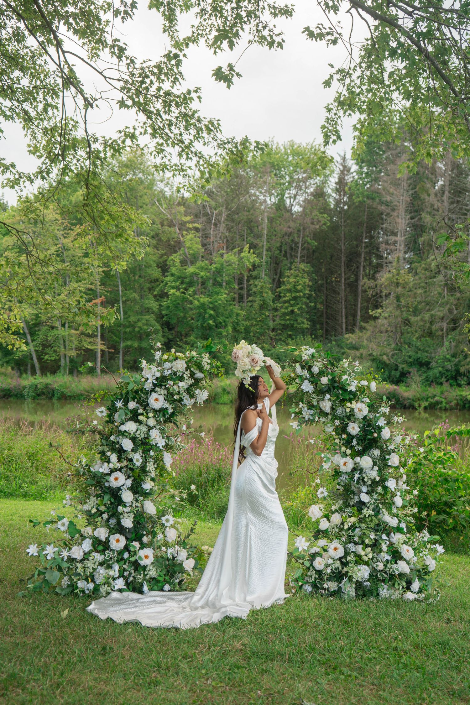 Bride in white gown stands before circular white floral arch, raising bouquet overhead near a woodland pond.