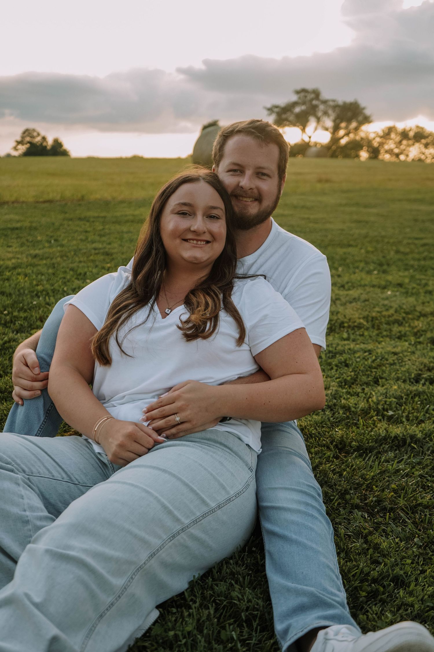 Smiling couple sitting together on lush green grass during a golden hour engagement photo session.
