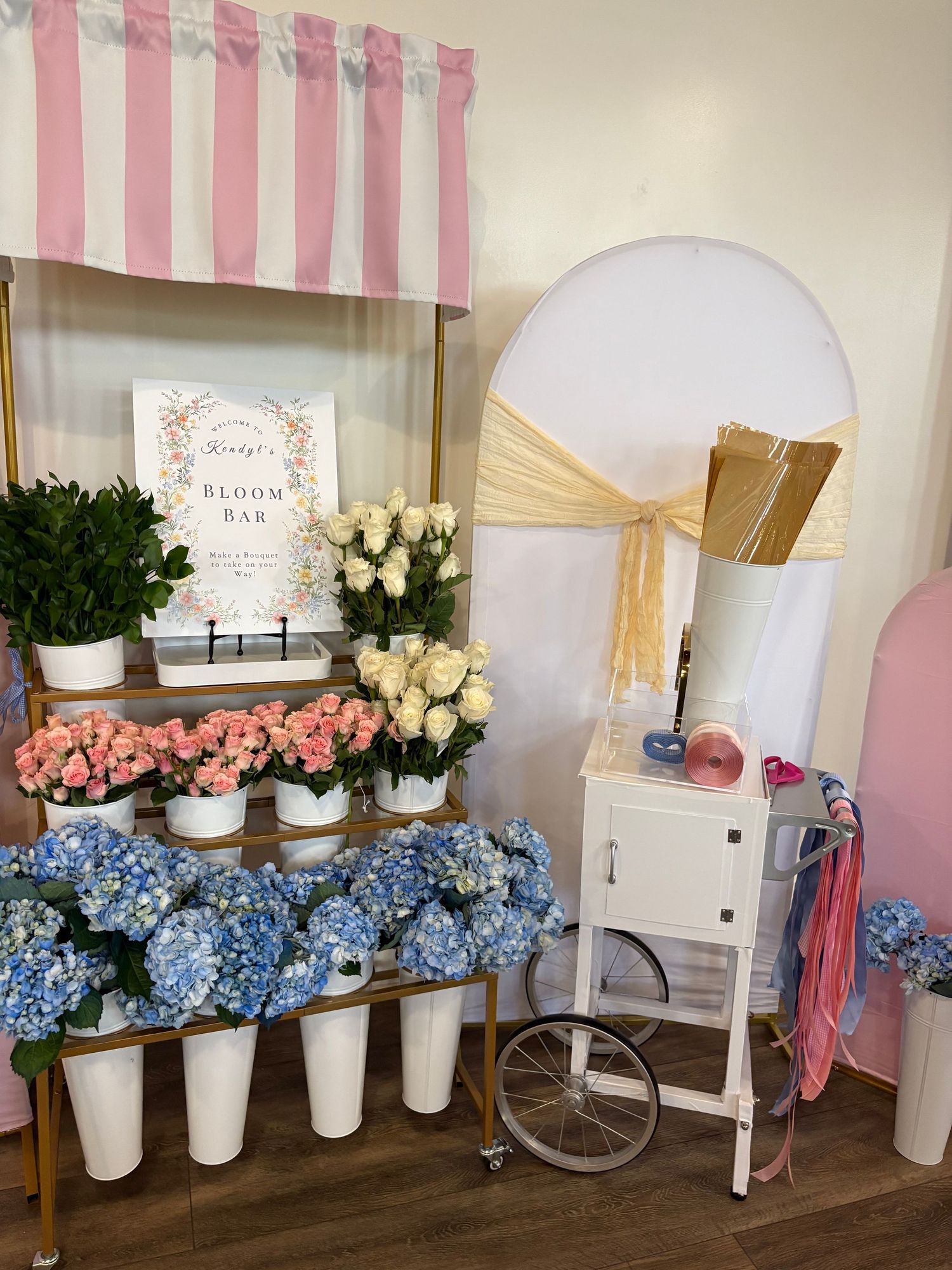 Elegant bloom bar display with pink and white striped canopy, featuring blue hydrangeas, pink and white roses in white vases.