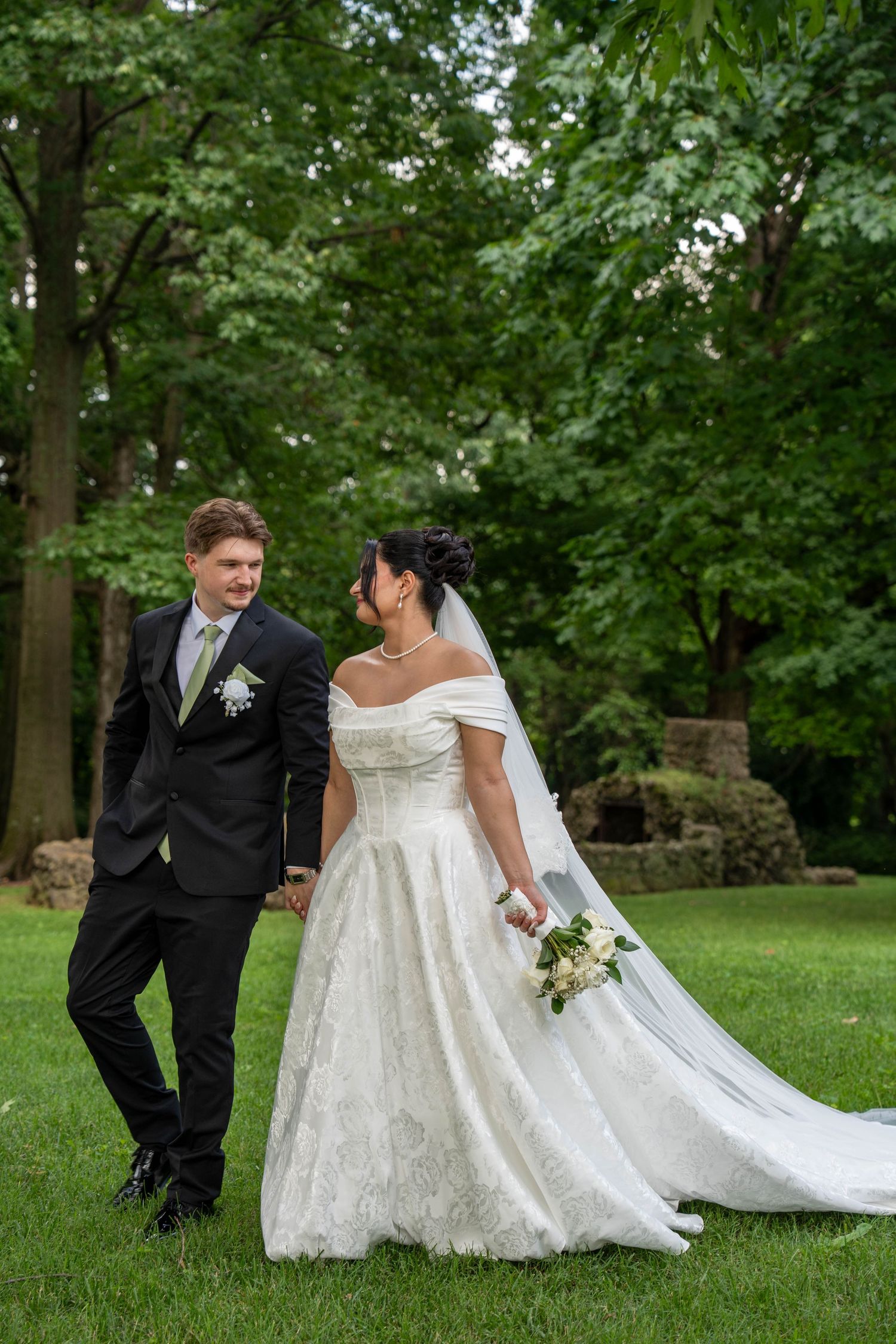 A bride and groom walk hand in hand on green grass surrounded by lush trees on their wedding day.