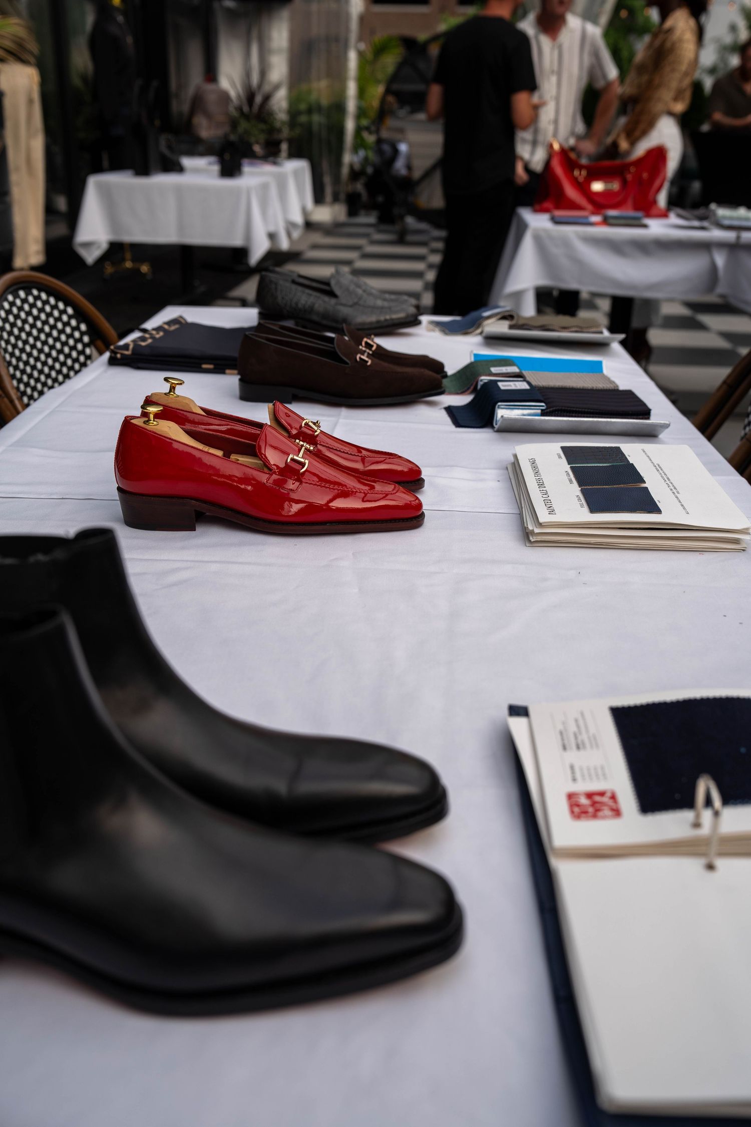 Display of luxury leather shoes including red loafers and black boots on a white table with fabric swatches at a fashion event.