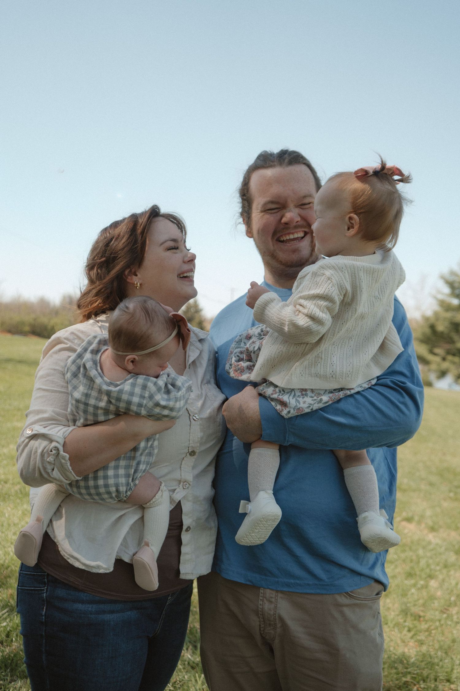 Happy family of four with parents holding twin babies outdoors in green field under blue sky.