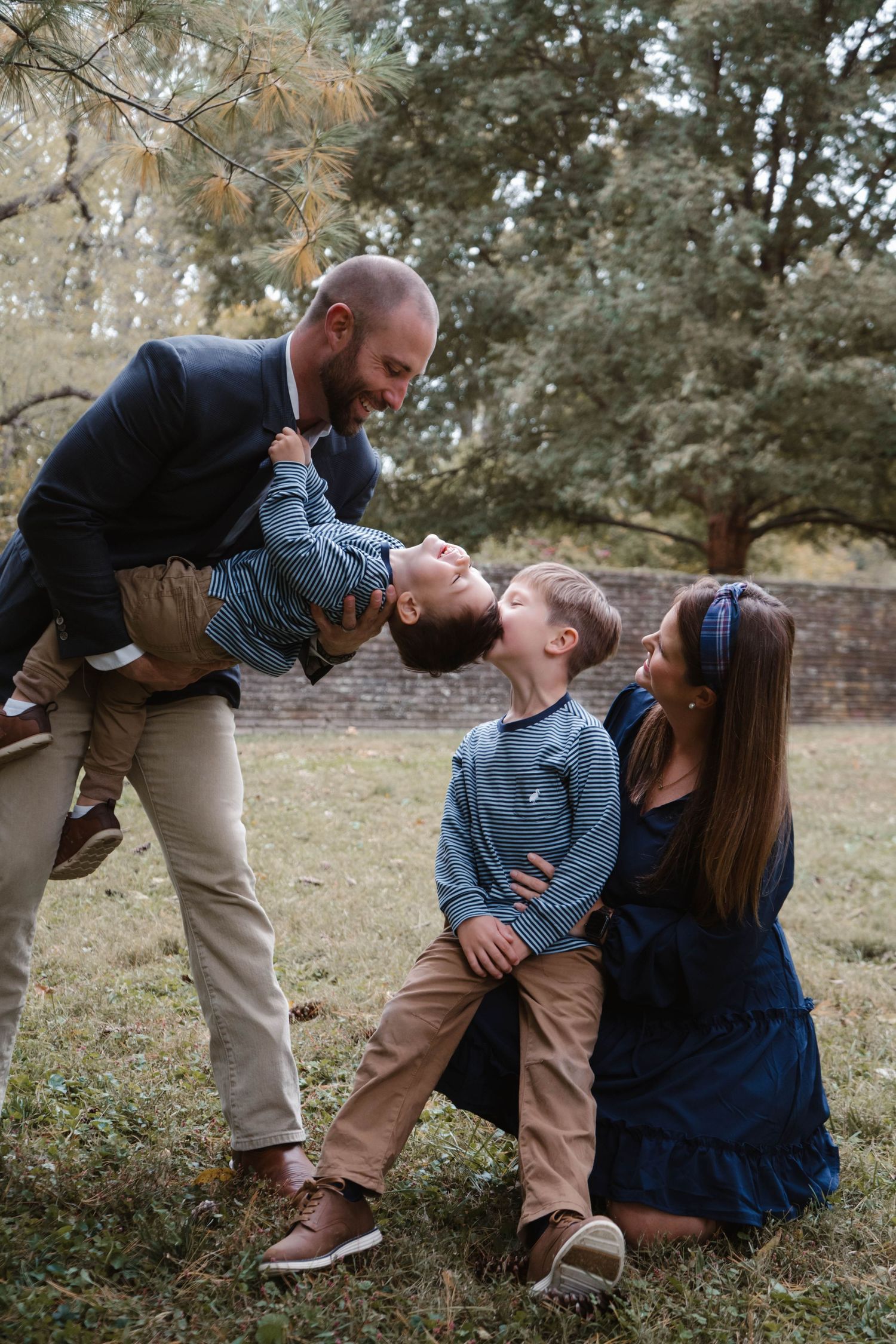 Playful family of four laughs together outdoors as father dips toddler toward older brother while mother kneels smiling.