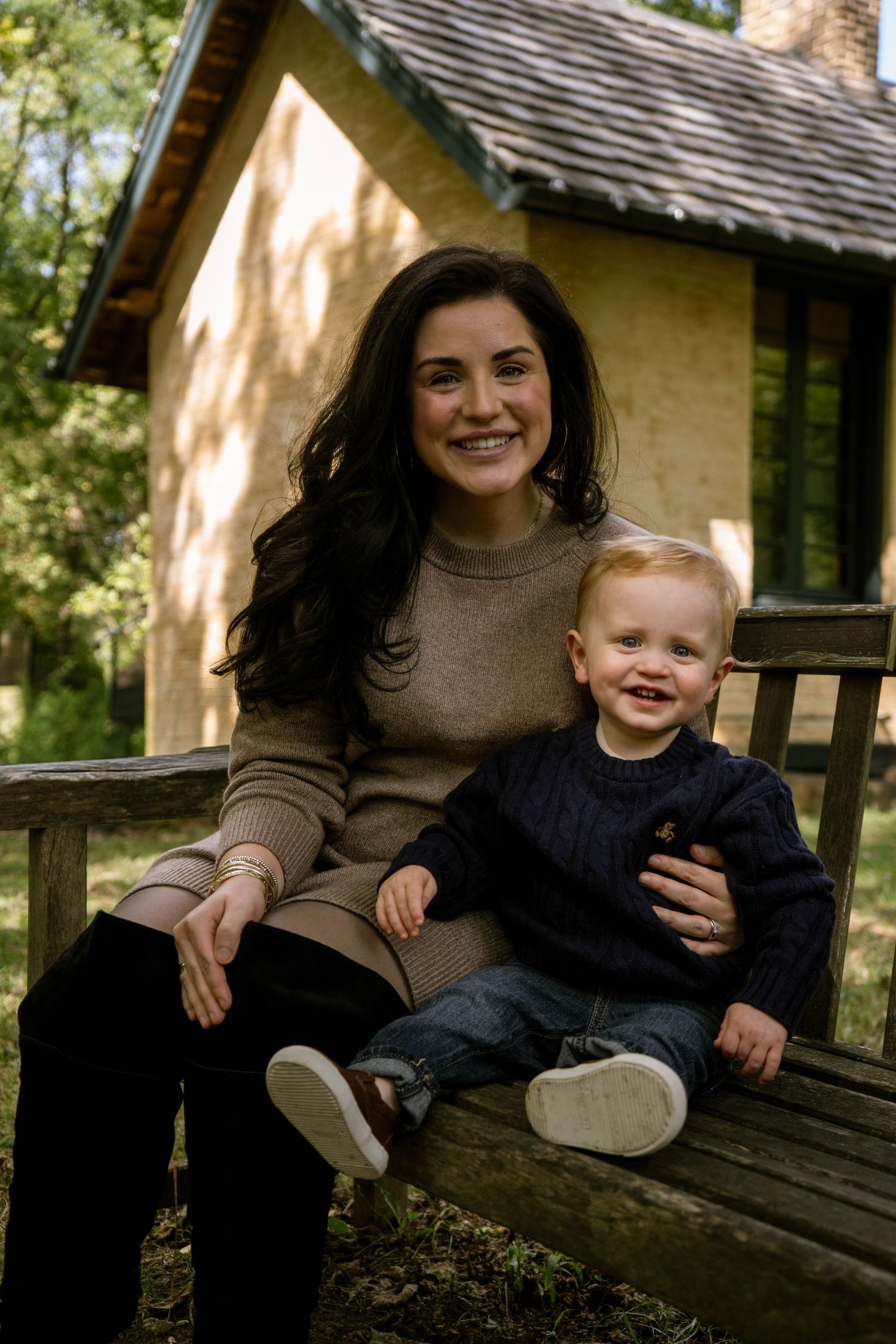 Smiling mother sits on wooden bench with happy toddler boy in front of a charming cottage.