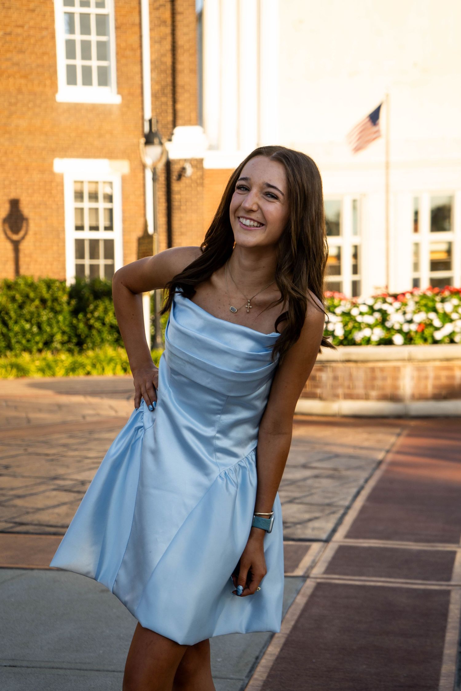 Laughing teenage girl in light blue strapless dress poses playfully outdoors in golden hour light.