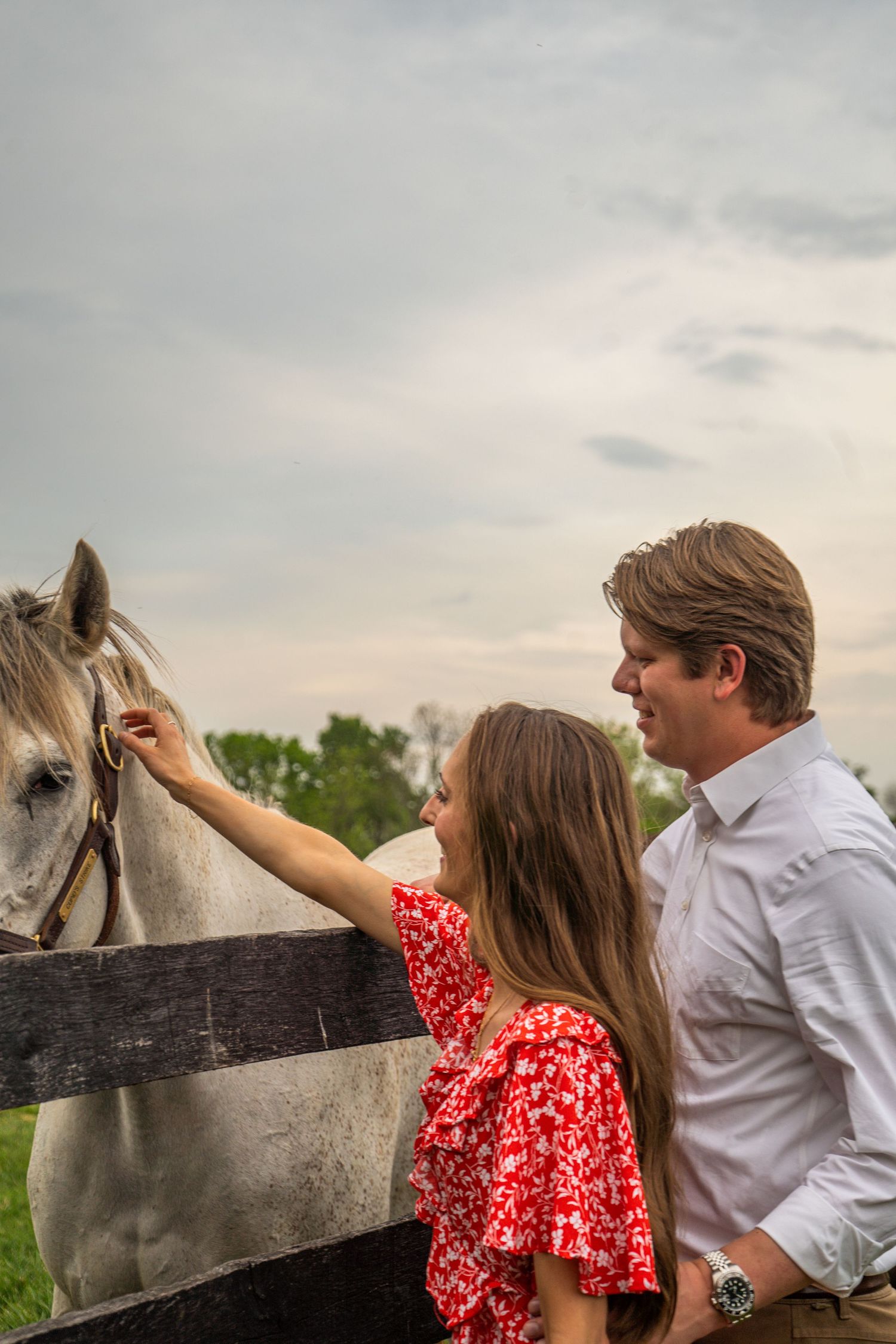 Engaged couple petting a white horse at a wooden fence on a farm under a cloudy sky.