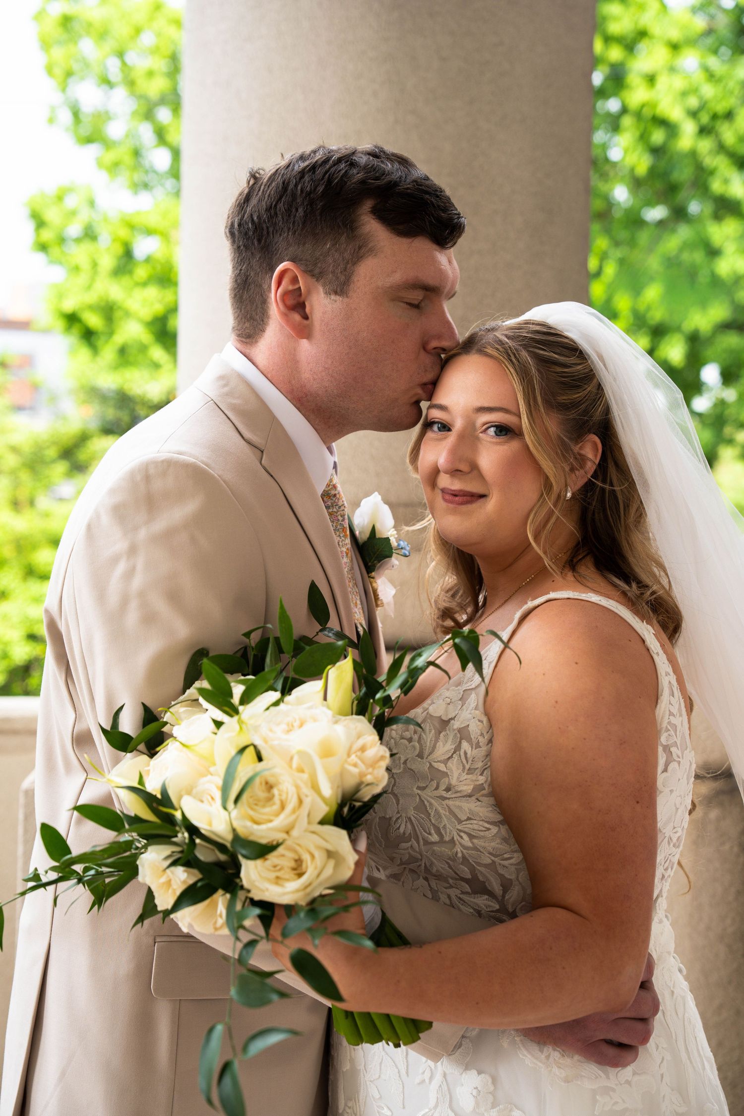 Groom in tan suit kisses bride on forehead as she smiles holding white rose wedding bouquet.