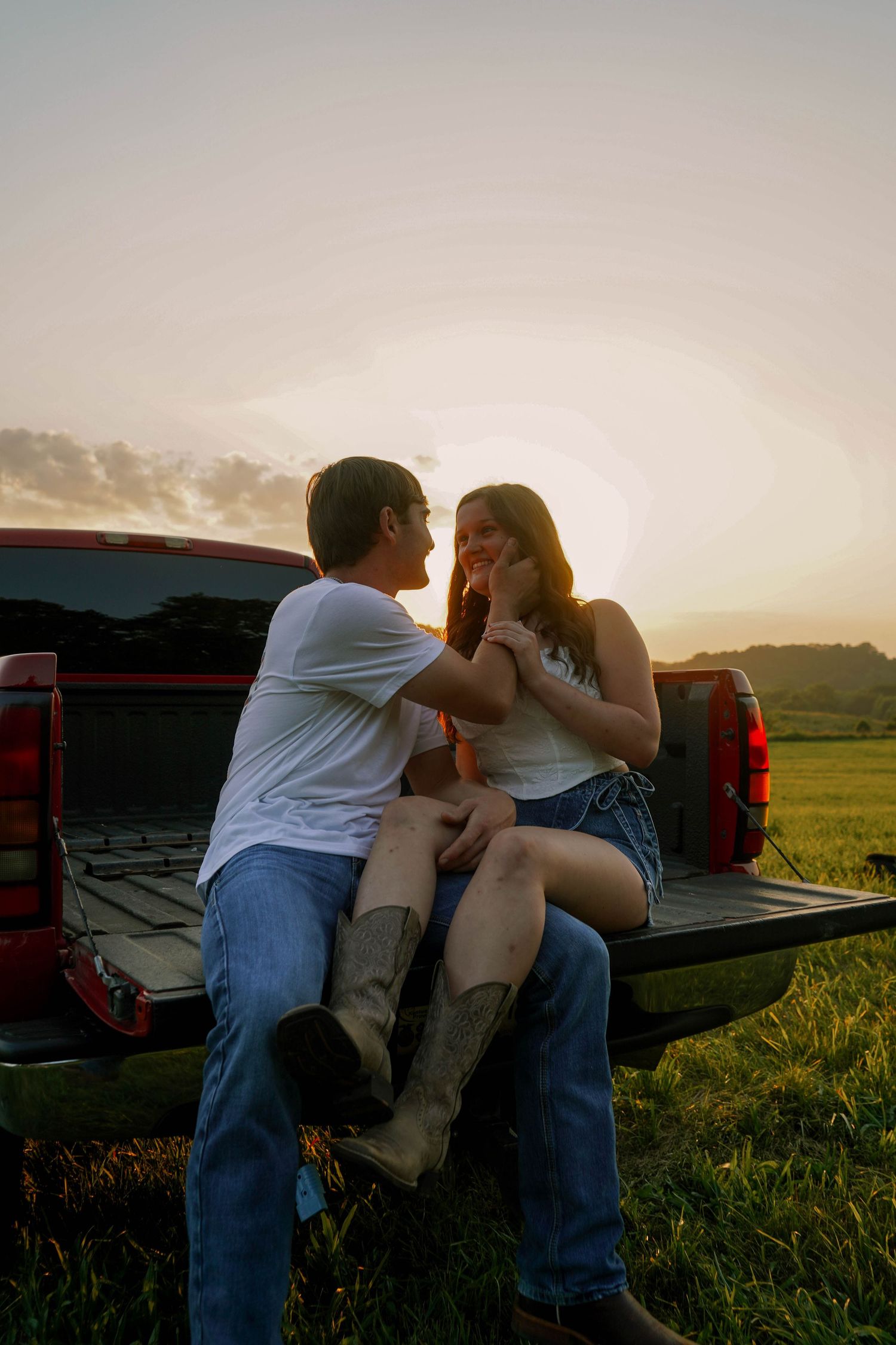 Romantic couple sitting on truck tailgate at sunset, man tenderly touching womans face in a scenic green meadow.