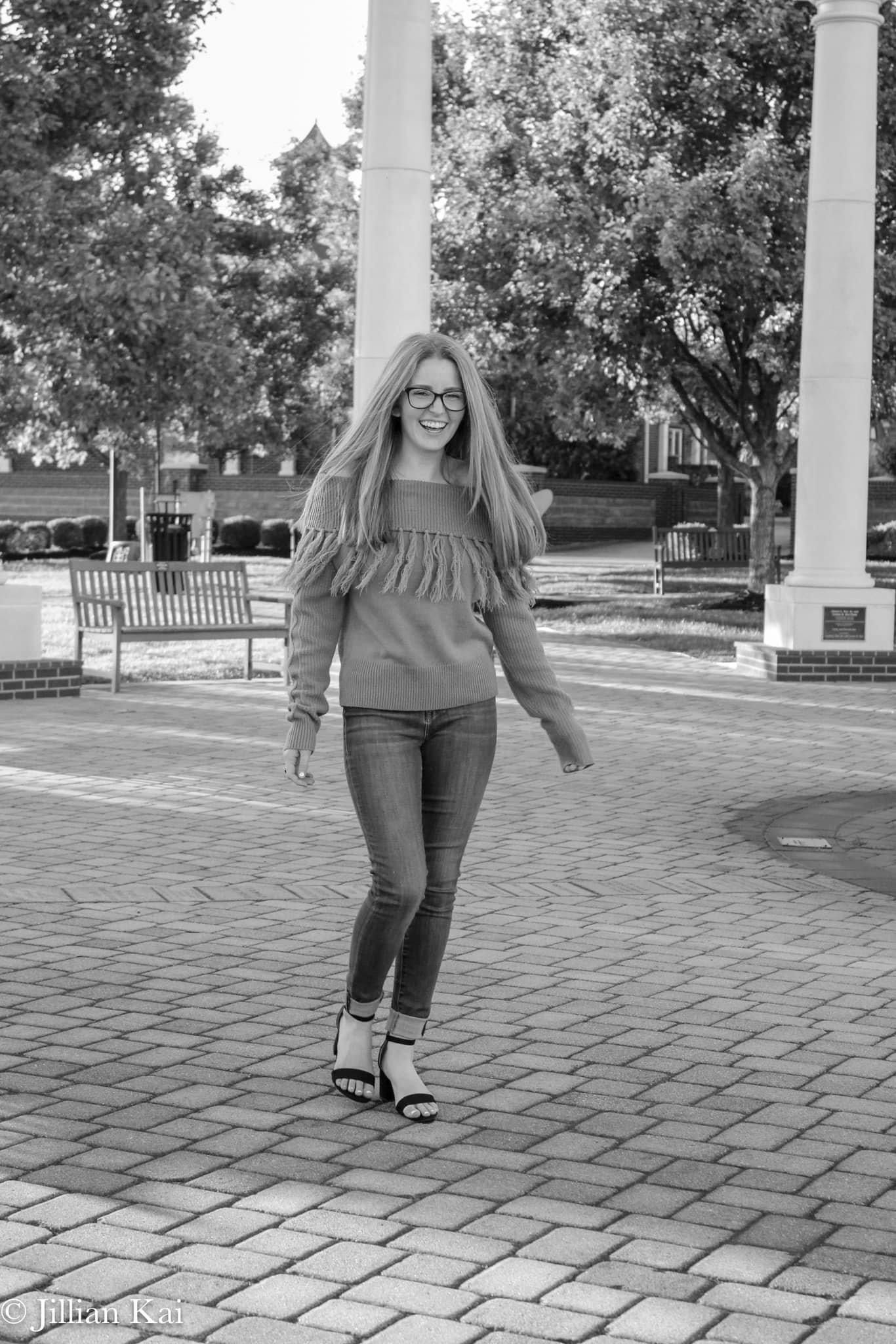 Young woman in fringe sweater and jeans smiles on brick plaza in black and white photo.