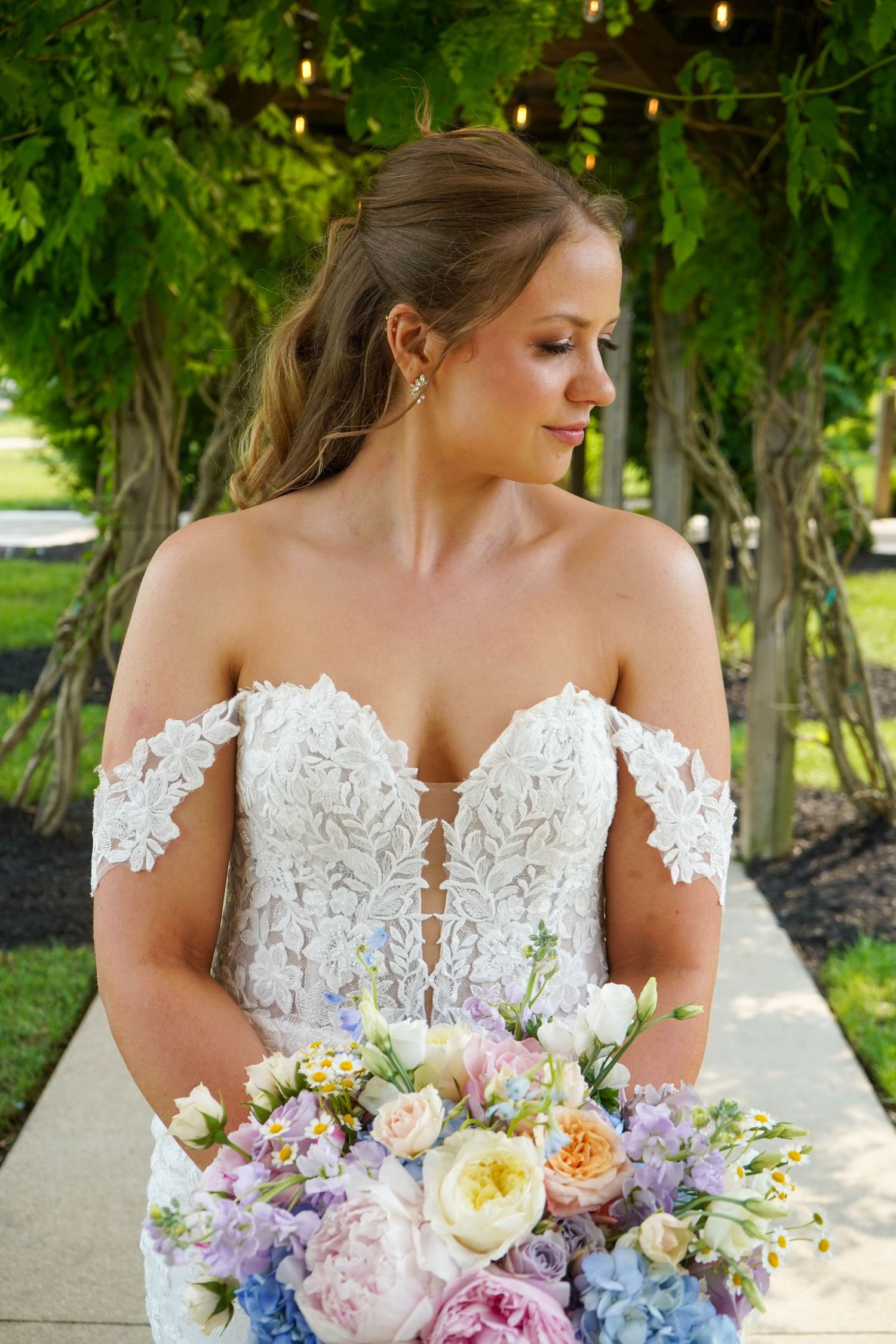 Bride in off-shoulder lace wedding gown holding pastel floral bouquet under green vine-covered arbor.