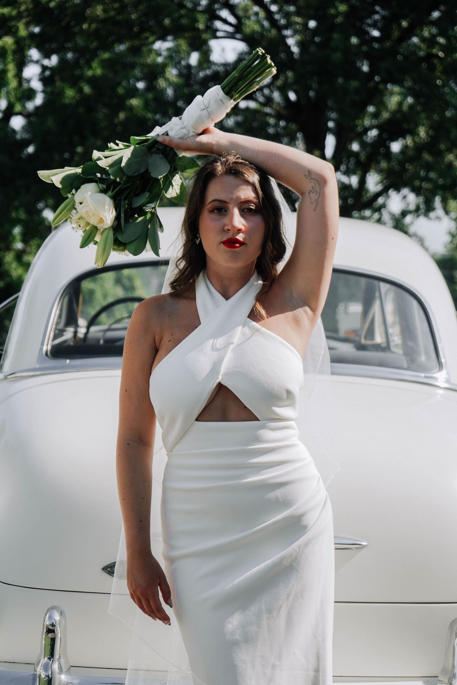 Elegant bride in white crossover halter dress raising bouquet overhead beside classic vintage white car.