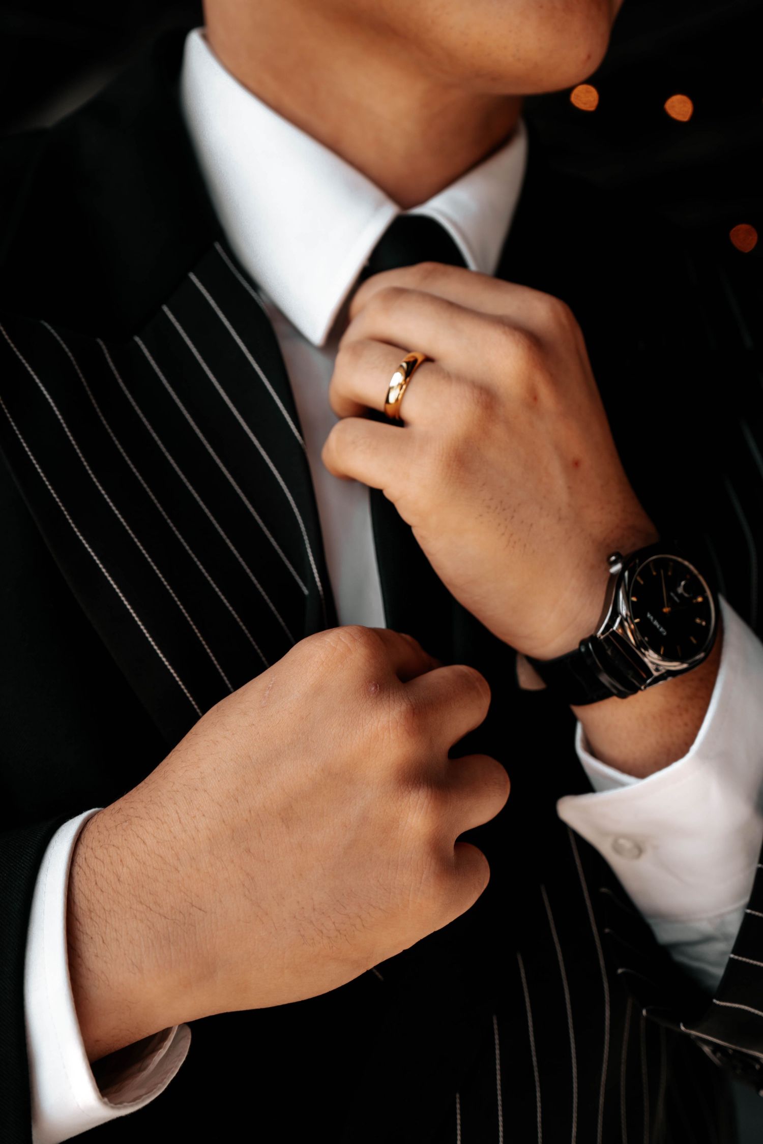 Close-up of groom in pinstripe suit adjusting black tie wearing gold wedding band and luxury watch.