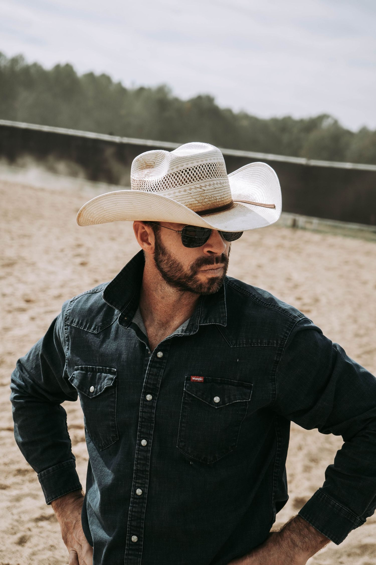 Rugged cowboy in white straw hat and sunglasses wearing black denim shirt outdoors at sandy ranch.