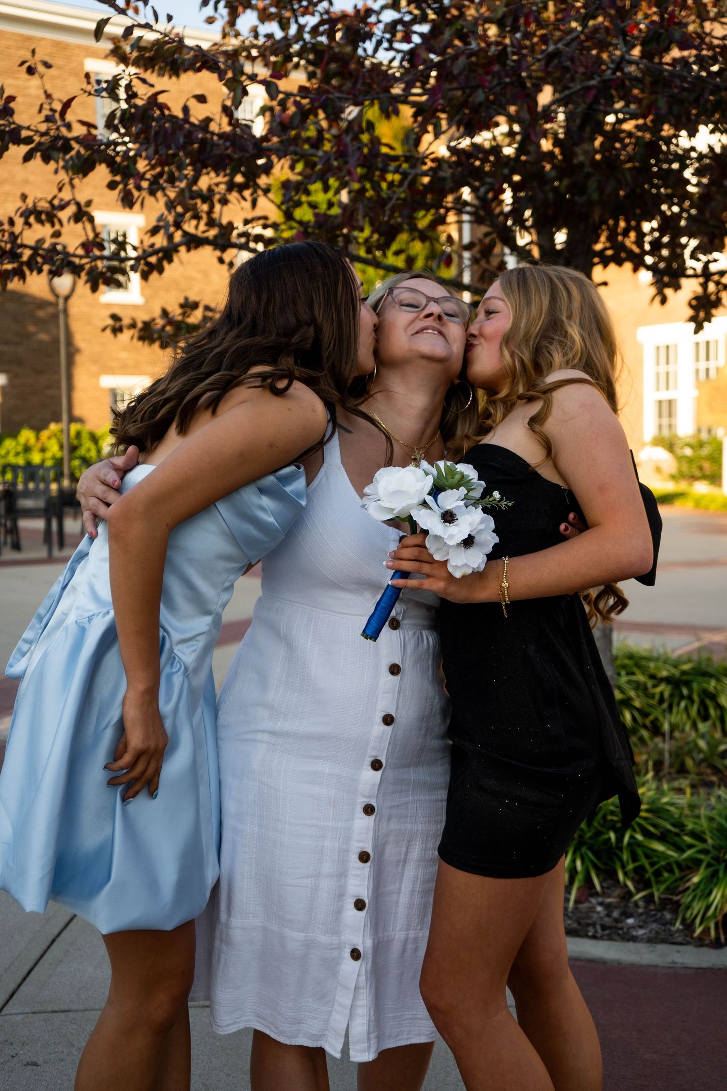 Three young women share a joyful moment as two kiss the cheeks of a woman in a white dress holding flowers.