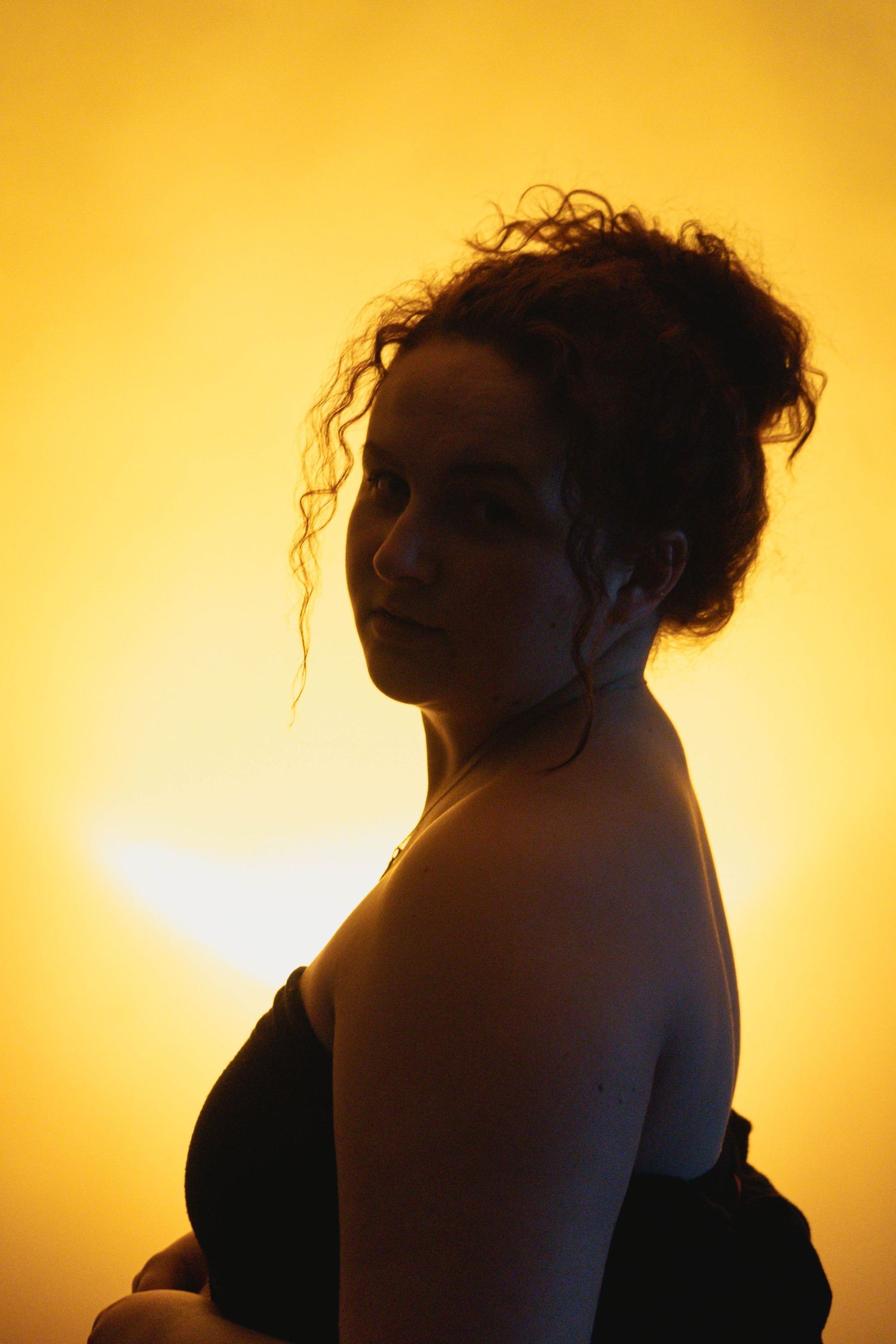 Side profile of curly-haired woman in black top glancing back against a glowing golden sunset background.