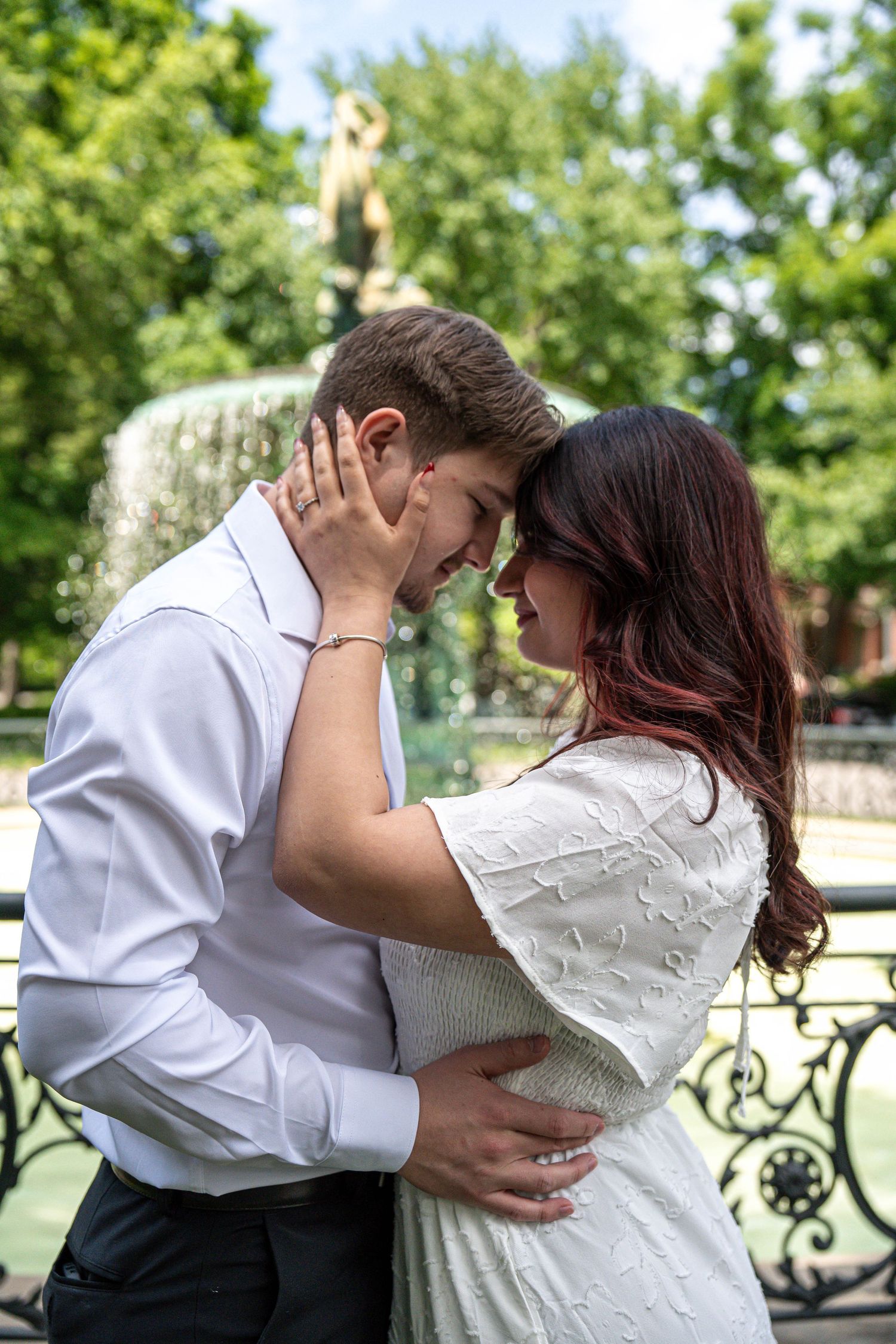 Young couple embracing and smiling at each other in a sunny park with a fountain and green trees behind them.