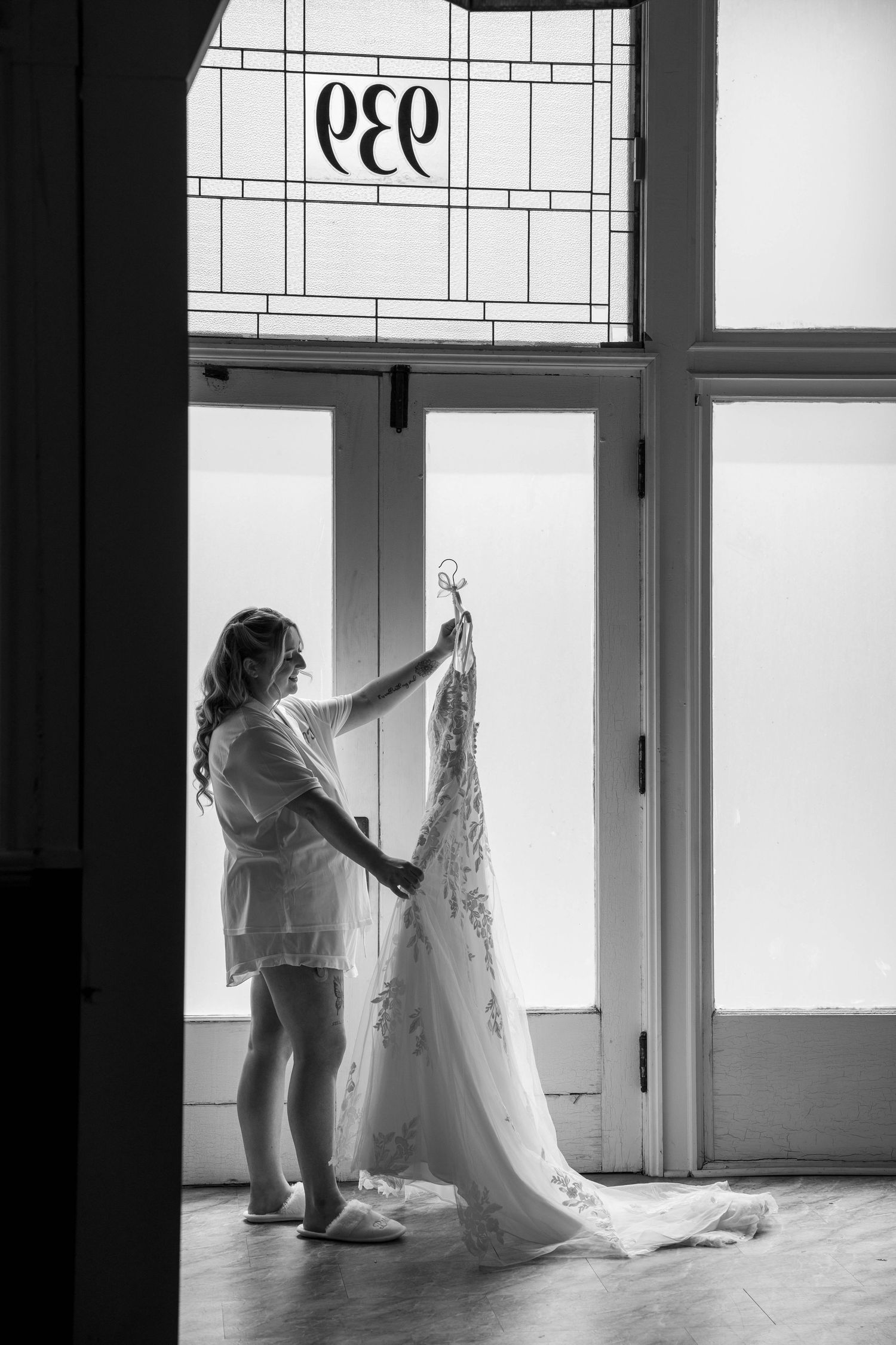Black and white photo of a bride holding her wedding dress up to a stained glass window.
