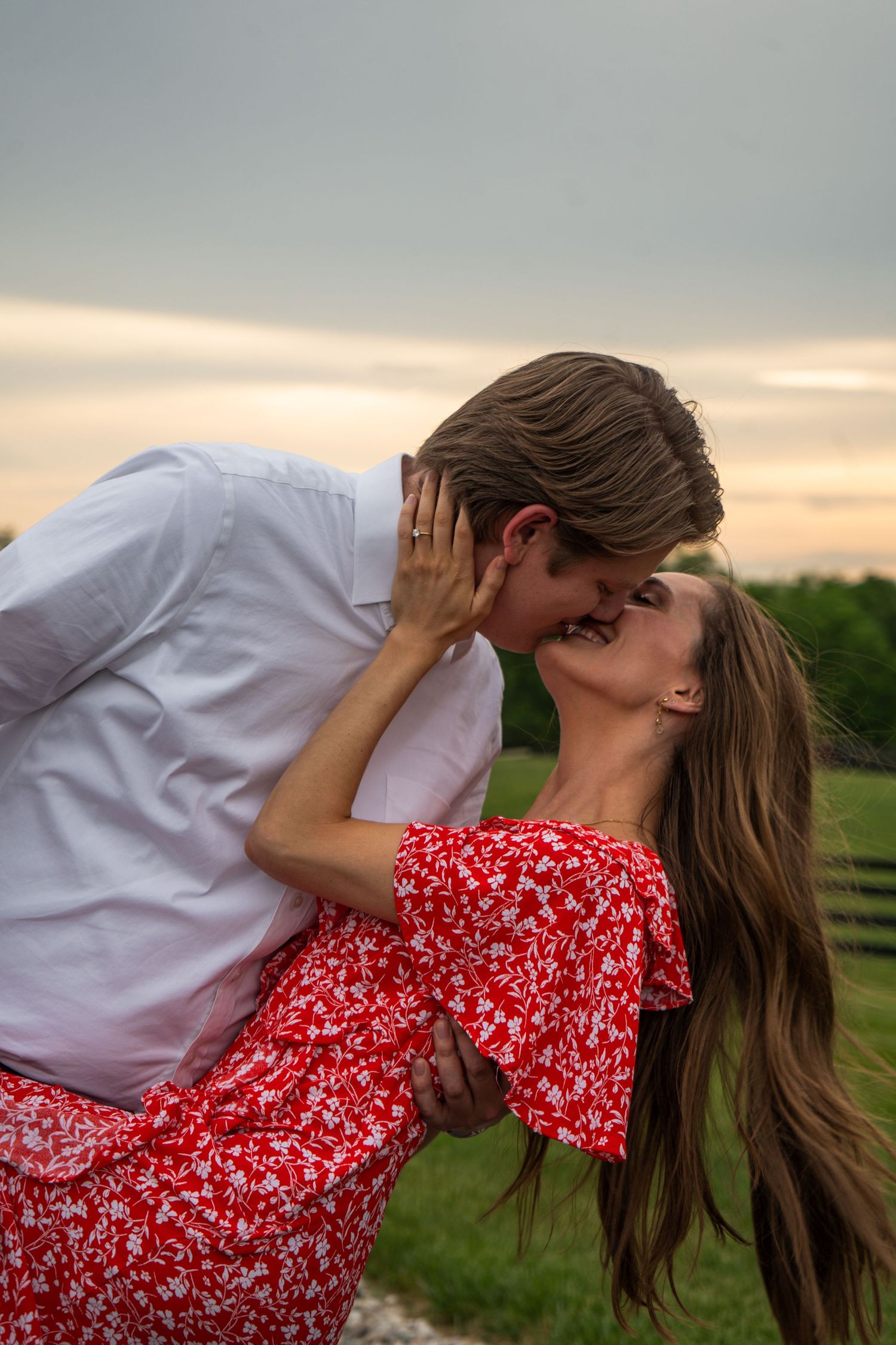 Couple sharing a passionate kiss outdoors at sunset with green farm fields in the background.