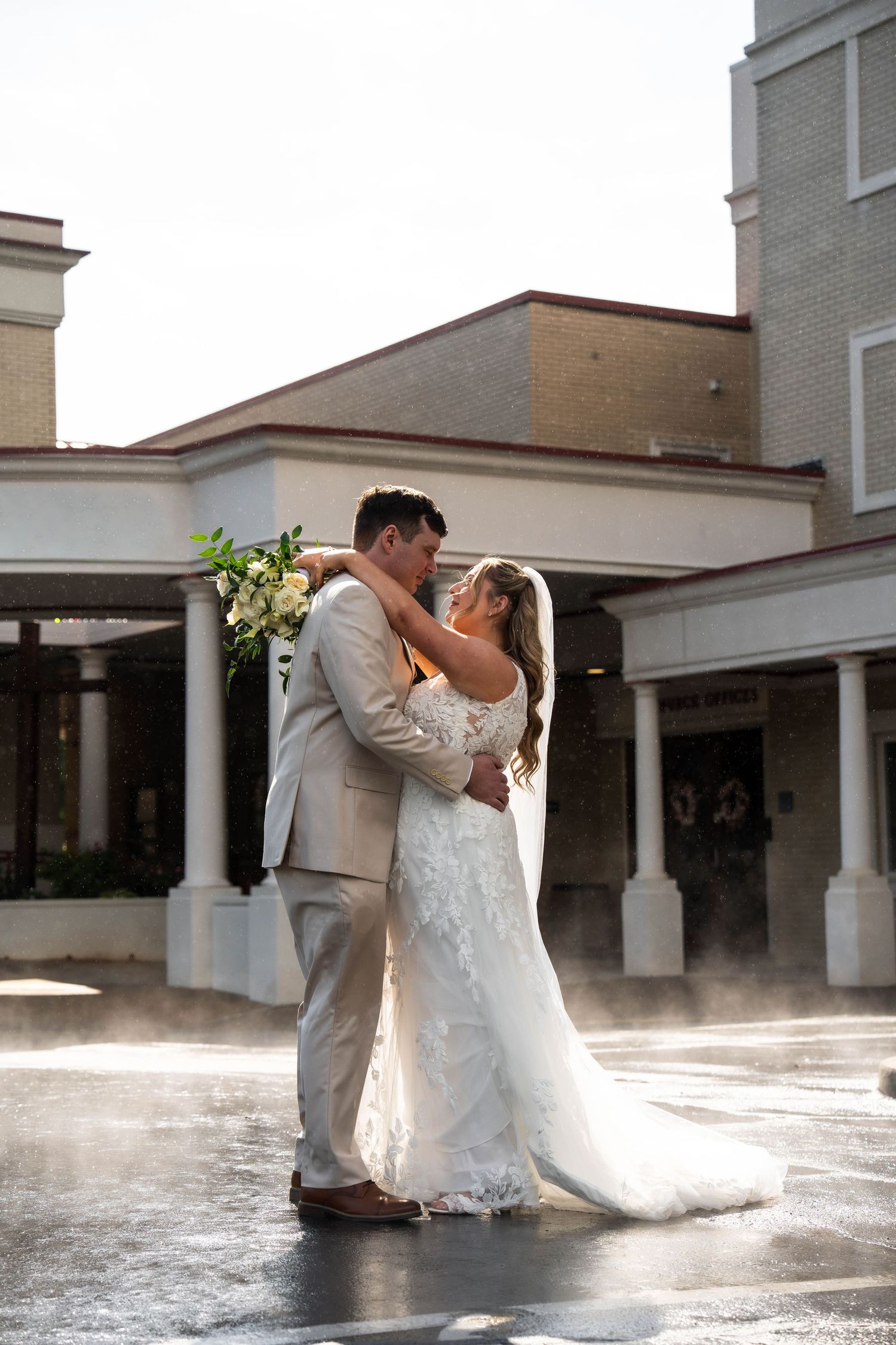Bride and groom embrace and nearly kiss in golden sunlight outside a classic building after wedding.