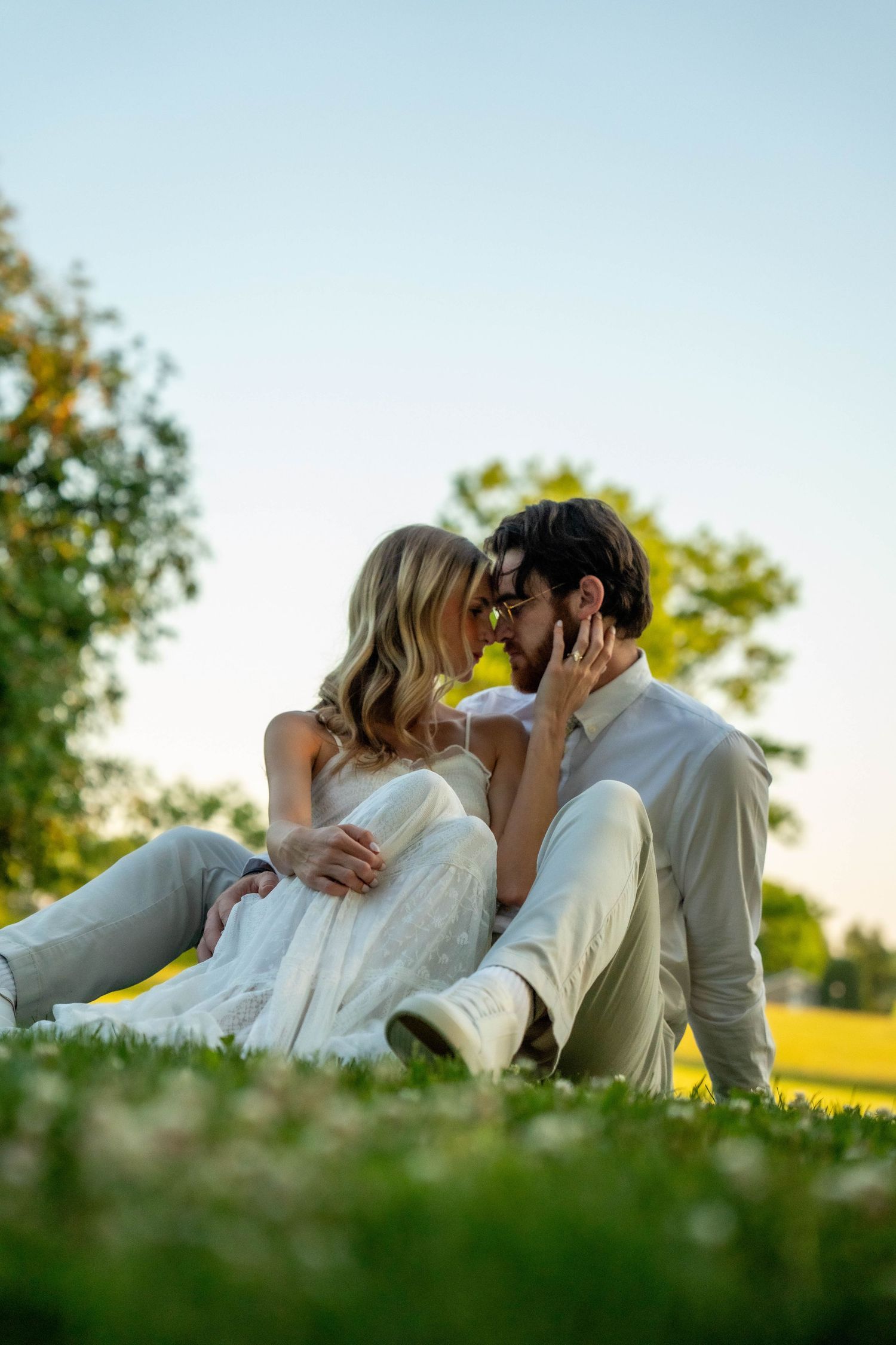 Bride and groom sit on grass in golden sunlight, leaning in for a kiss in a park.