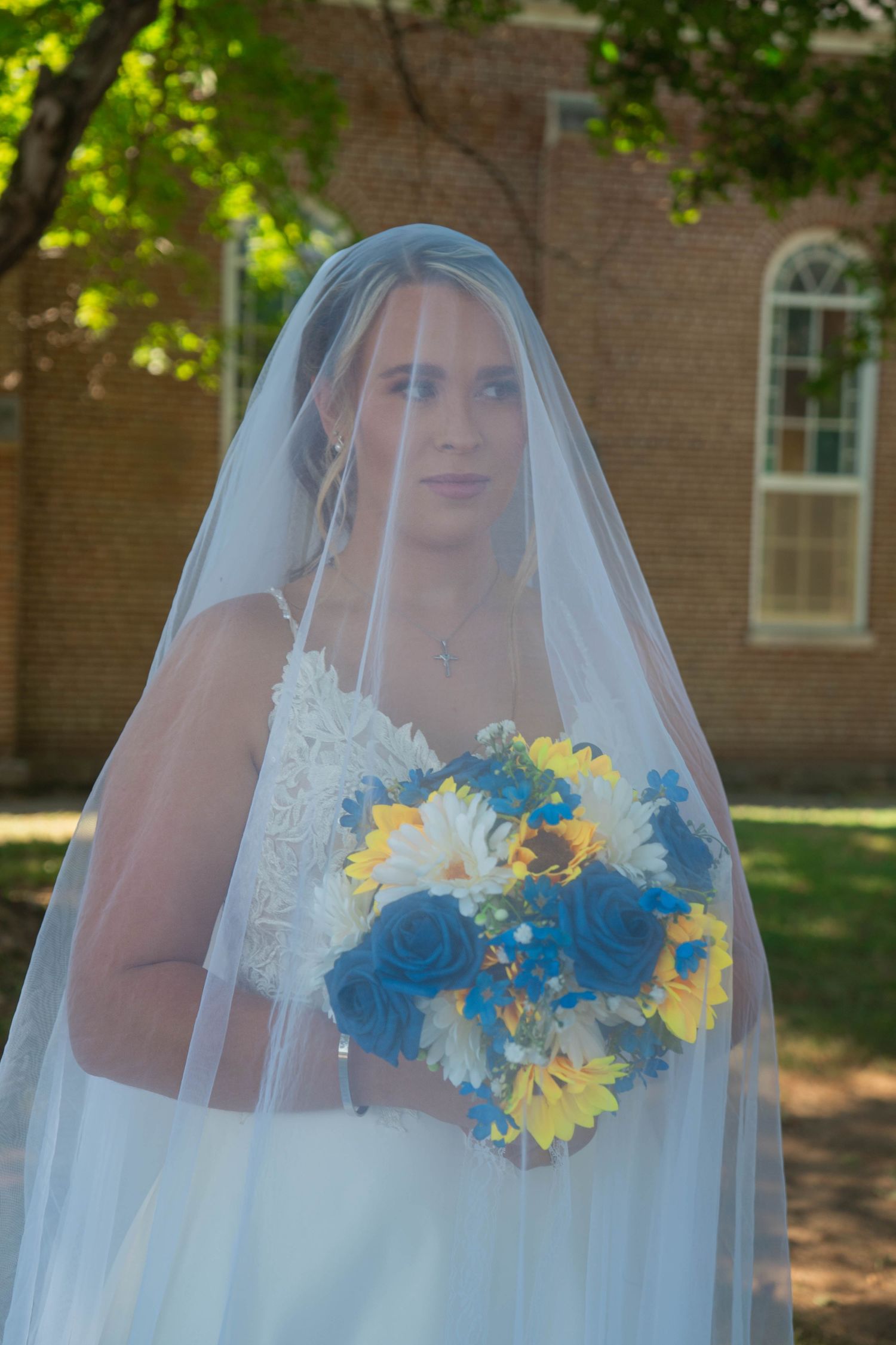 Bride wearing lace wedding gown and long veil holds blue and yellow floral bouquet outdoors near brick building.