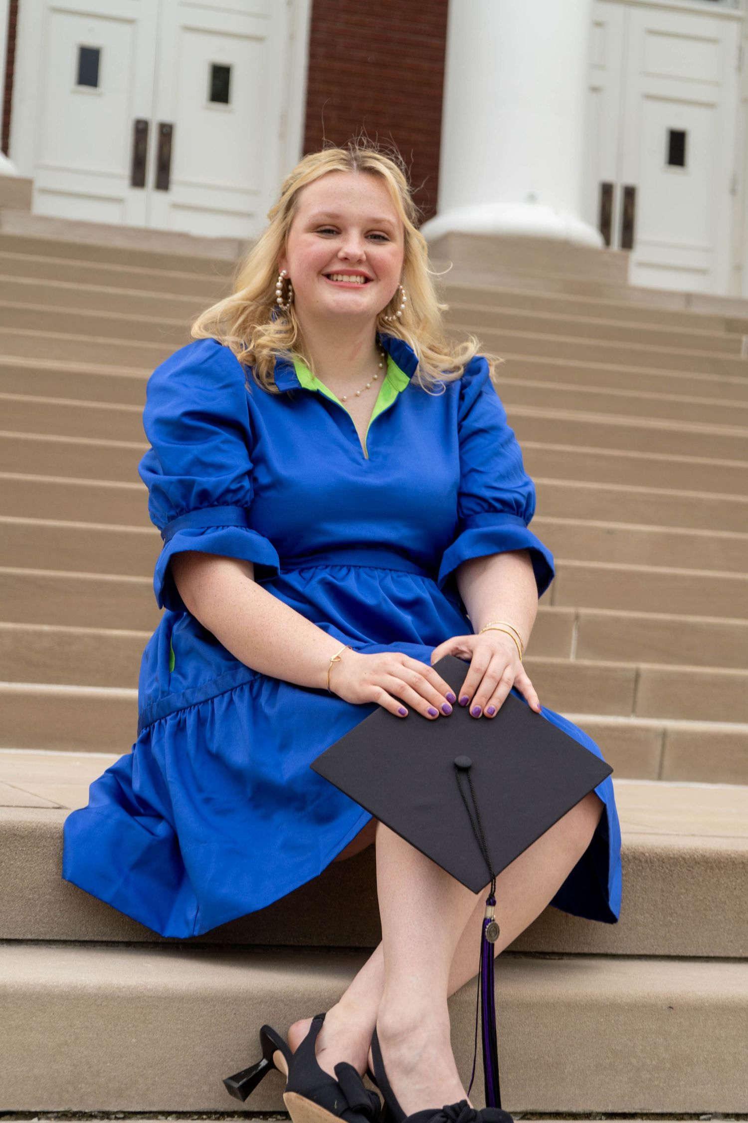 Smiling blonde graduate in blue dress sits on steps holding black mortarboard cap.