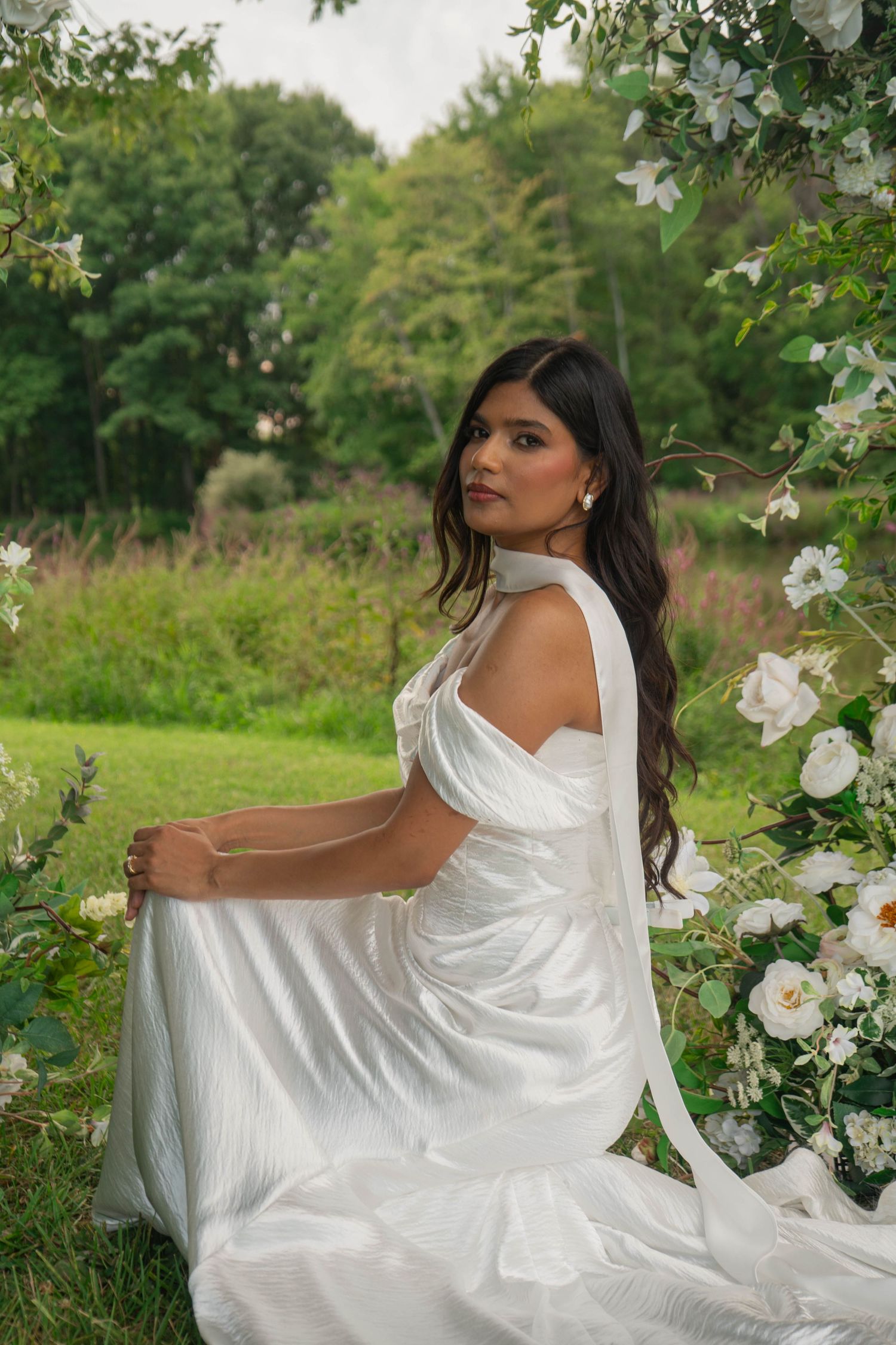 Bride in white satin gown sits gracefully beside white floral arch, looking back over her shoulder outdoors.