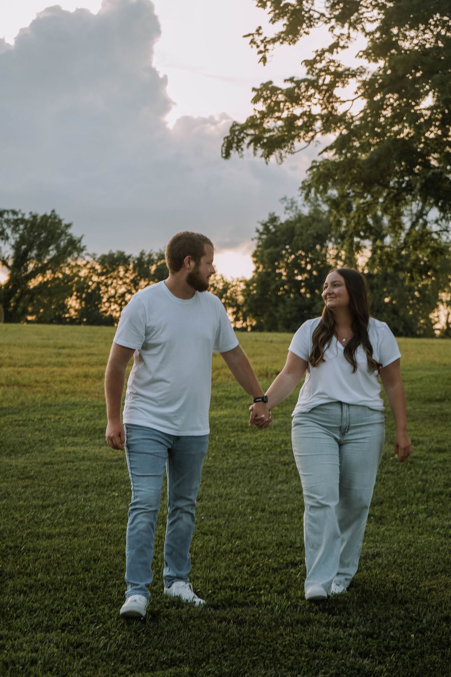 Happy couple in matching white tees and jeans holding hands walking through a green field at sunset.