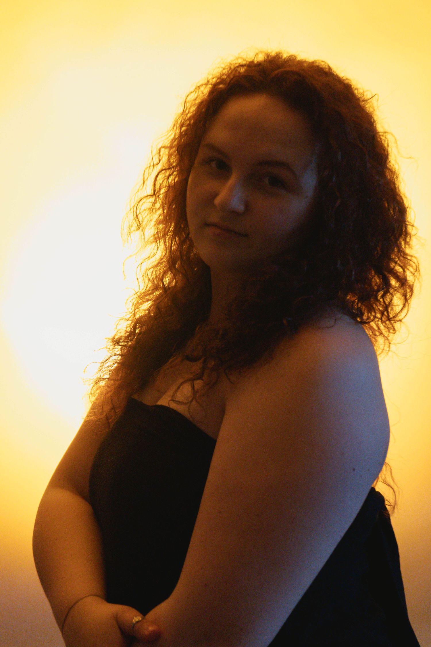 Young curly-haired woman in black top posing in side profile against glowing golden yellow backdrop.