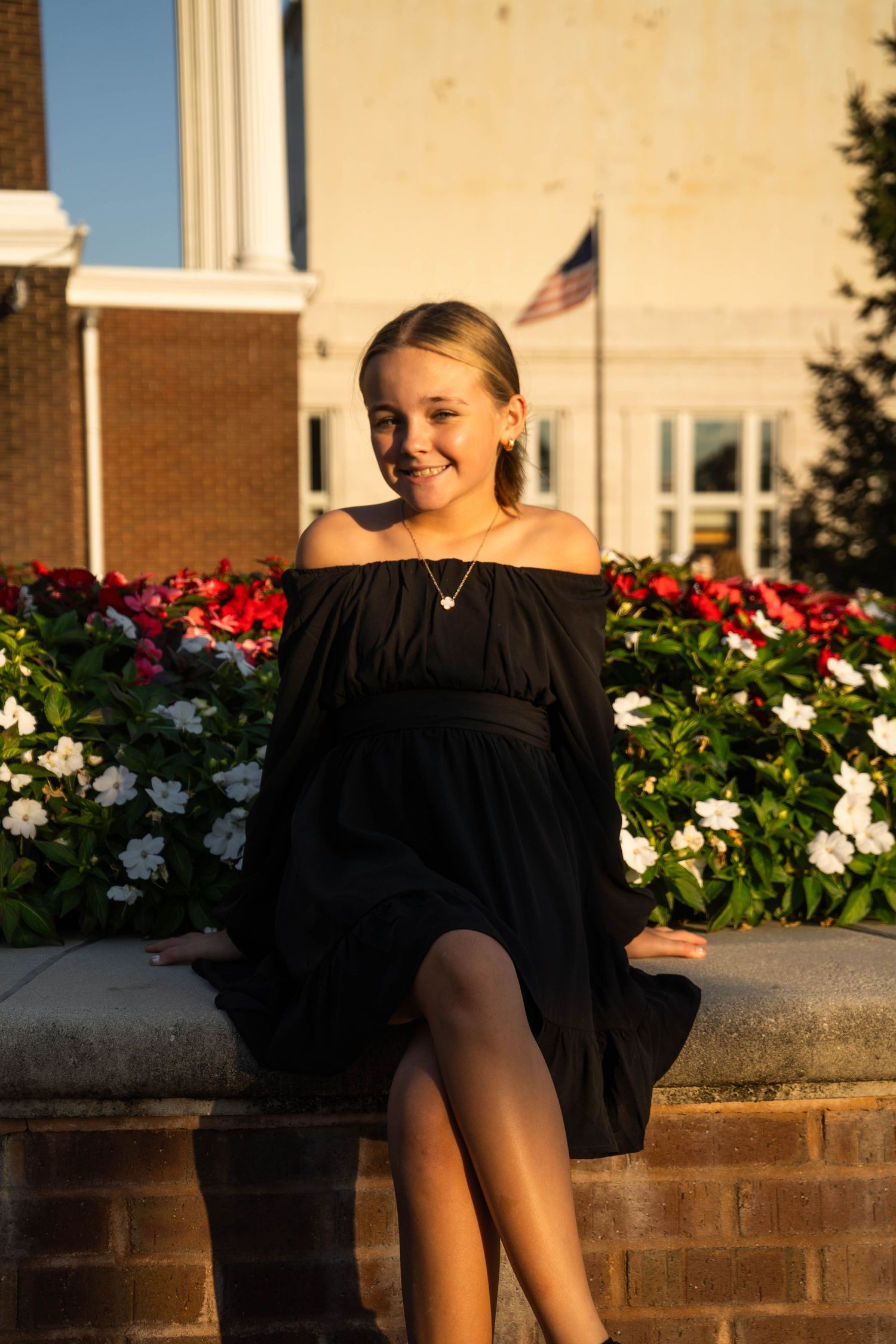 A smiling blonde teen girl in an off-shoulder black dress sits by red and white flowers outdoors.