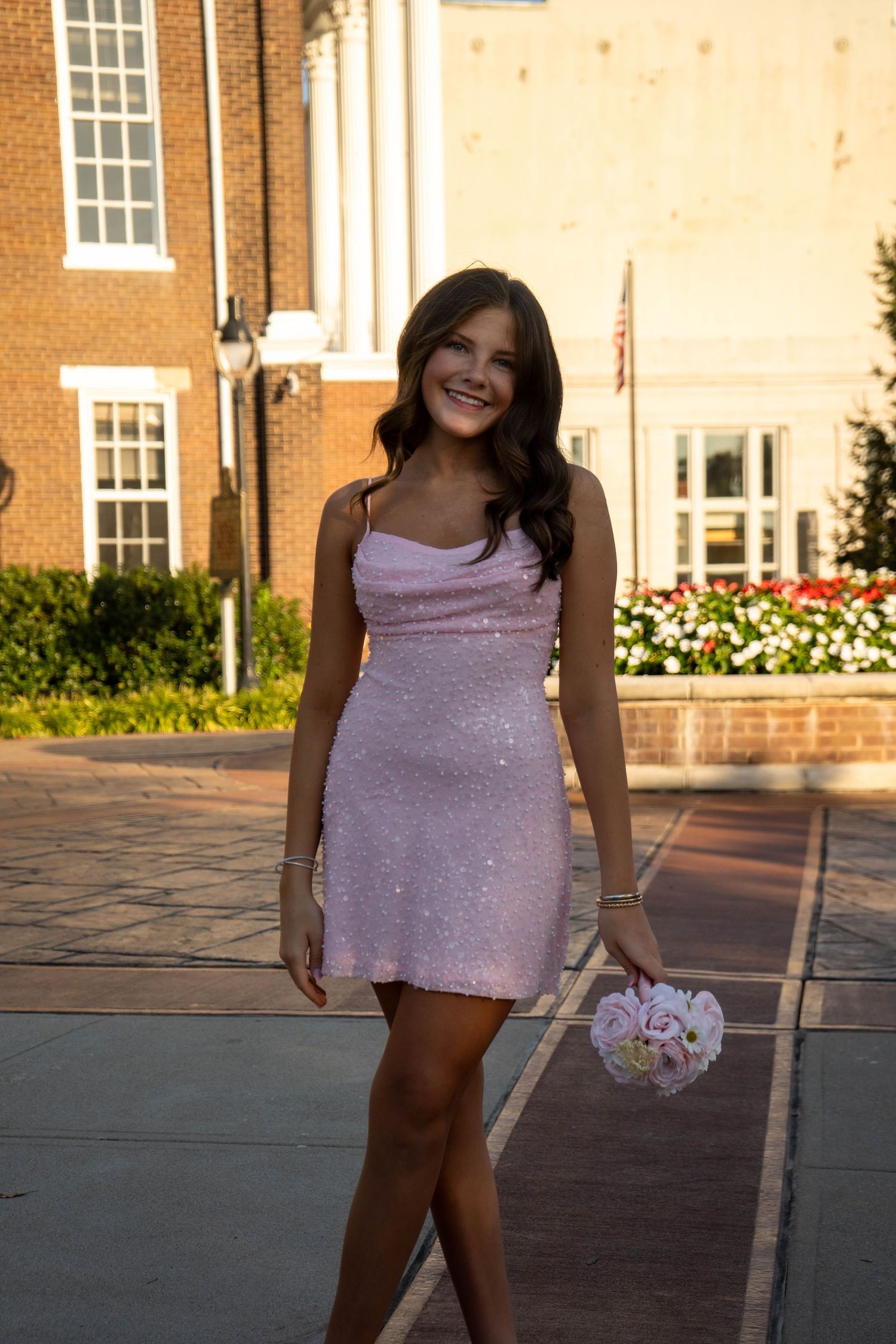 Young woman in pink sequin mini dress holding floral bouquet poses outdoors near brick building.